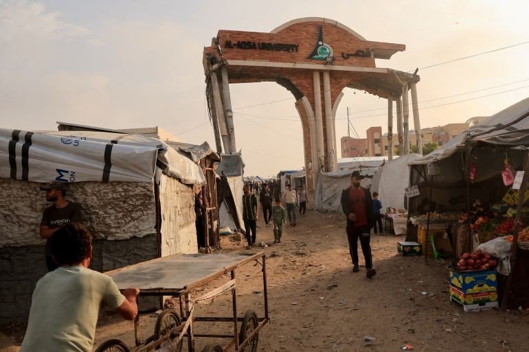 People walk through the damaged gate of Al-Aqsa University surrounded by tents sheltering displaced Palestinians, in Khan Younis, southern Gaza, on November 24, 2025 