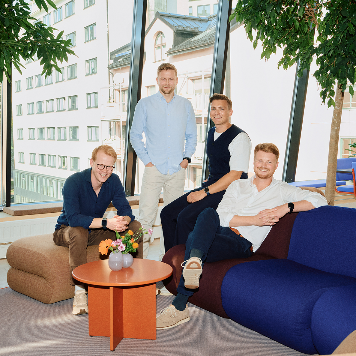 Fire menn smiler og sitter i et lyst lokale med store vinduer.