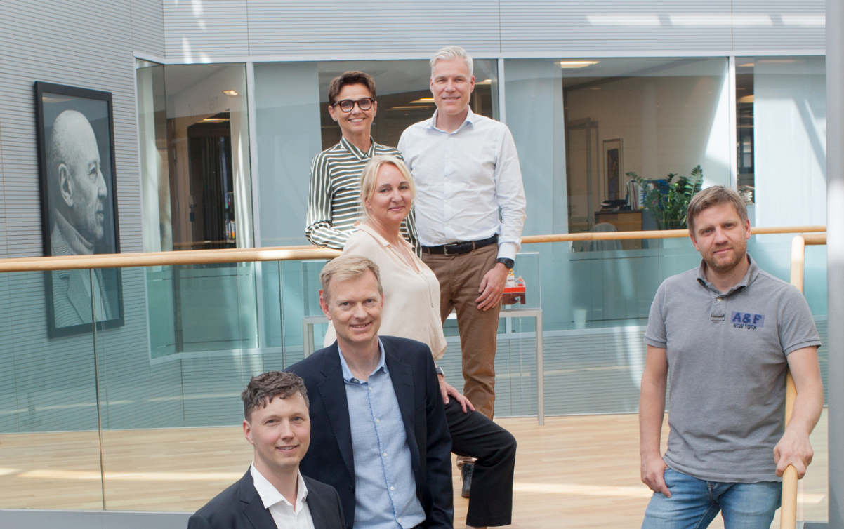 Fem personer smiler til kameraet i en lys kontorbygning, med en svart-hvitt-portrett i bakgrunnen.