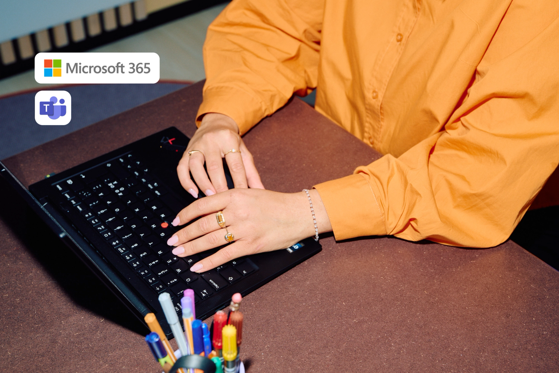 Kvinne i oransje skjorte med gullringer på fingrene jobber på en bærbar PC med Microsoft 365 og Teams-logoer.