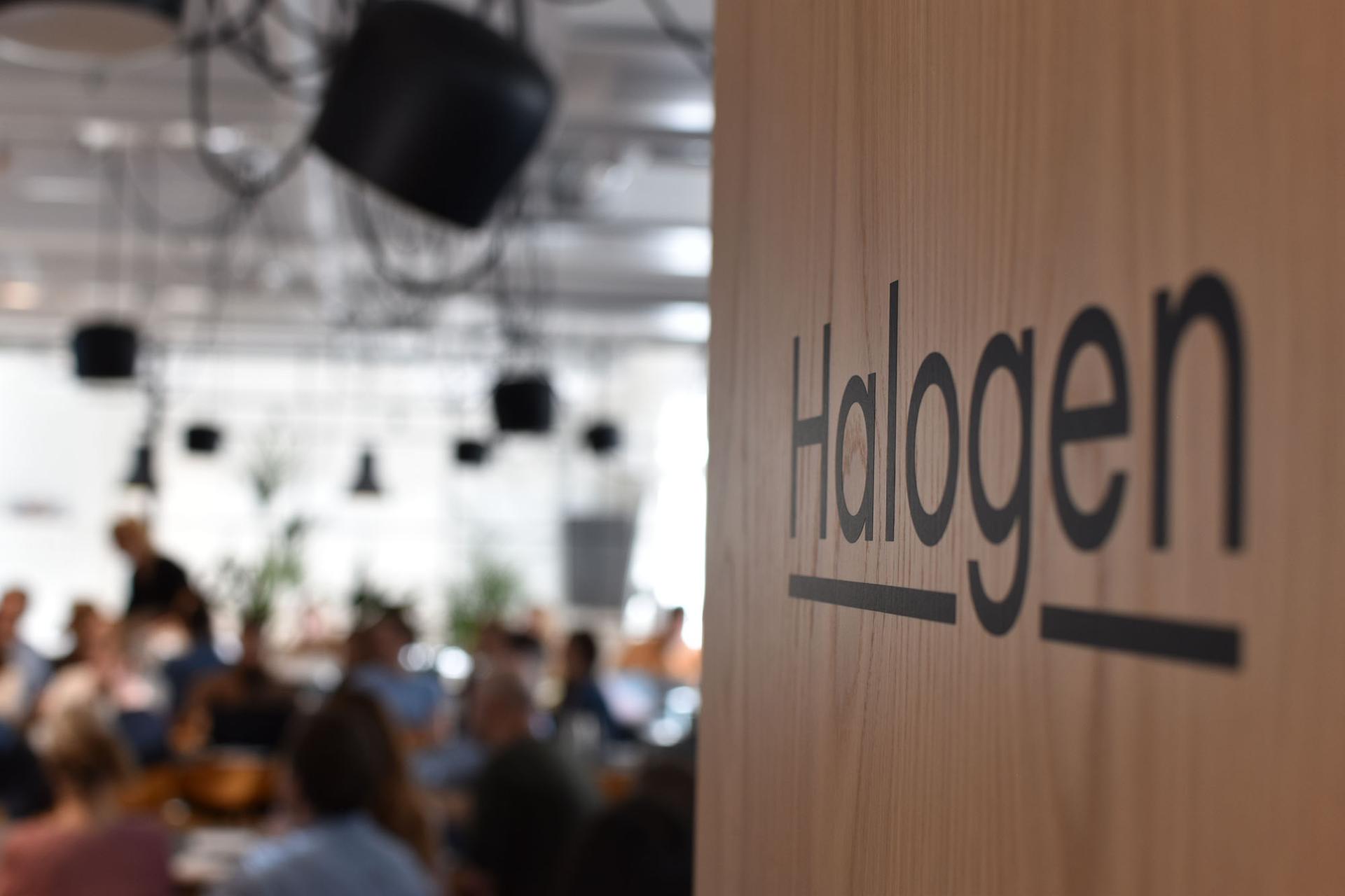 Bilde av Halogens kontorlokaler med teksten "Halogen" på en trevegg i forgrunnen og utydelige personer i bakgrunnen.