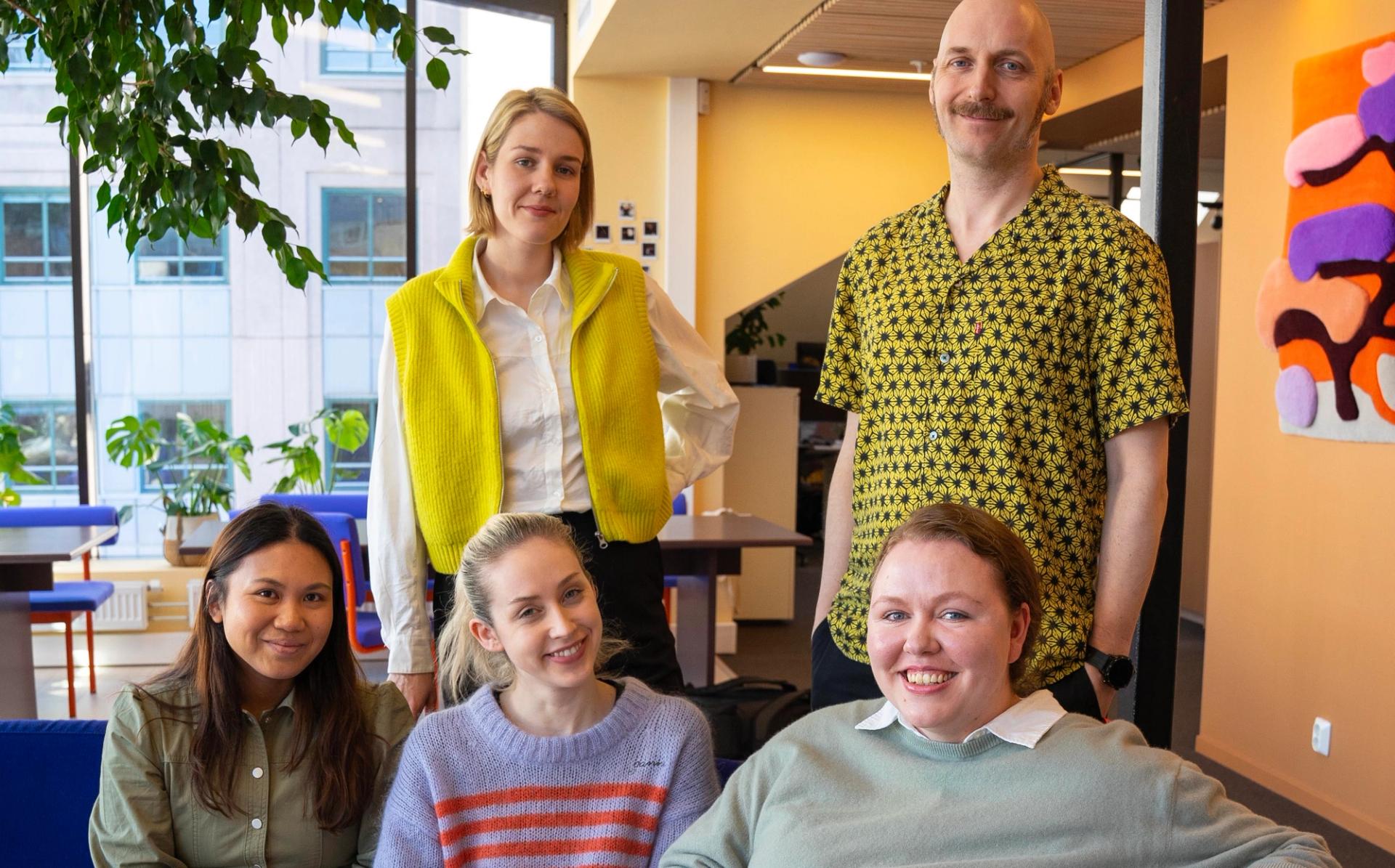 Fem smilende mennesker poserer innendørs foran et stort vindu og en vegg med kunst.