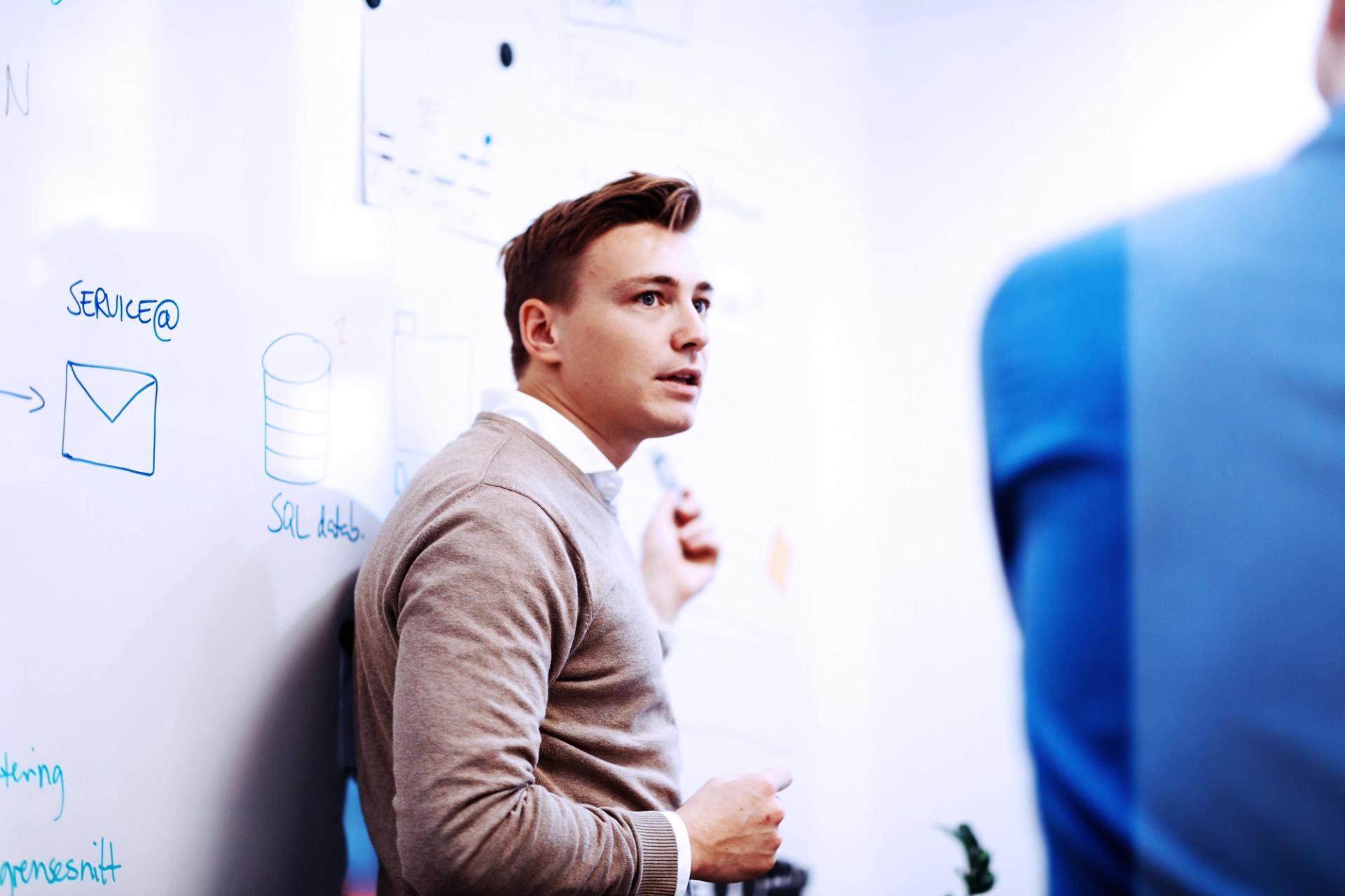 Ung mann presenterer ideer på en hvit tavle med diagrammer, mens en annen person lytter.