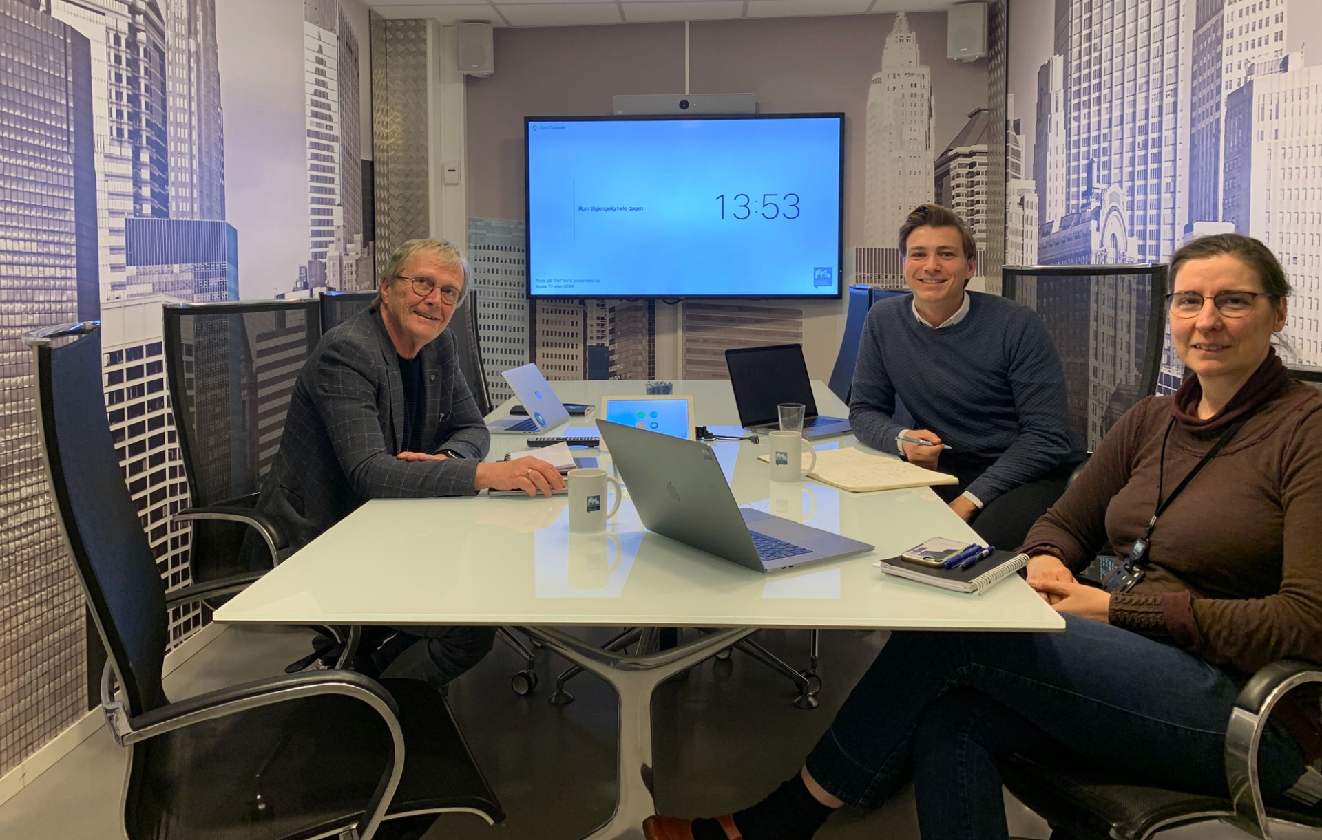 Tre personer smiler og sitter rundt et møtebord med laptoper og kaffekopper, foran en vegg med bilde av skyskrapere.
