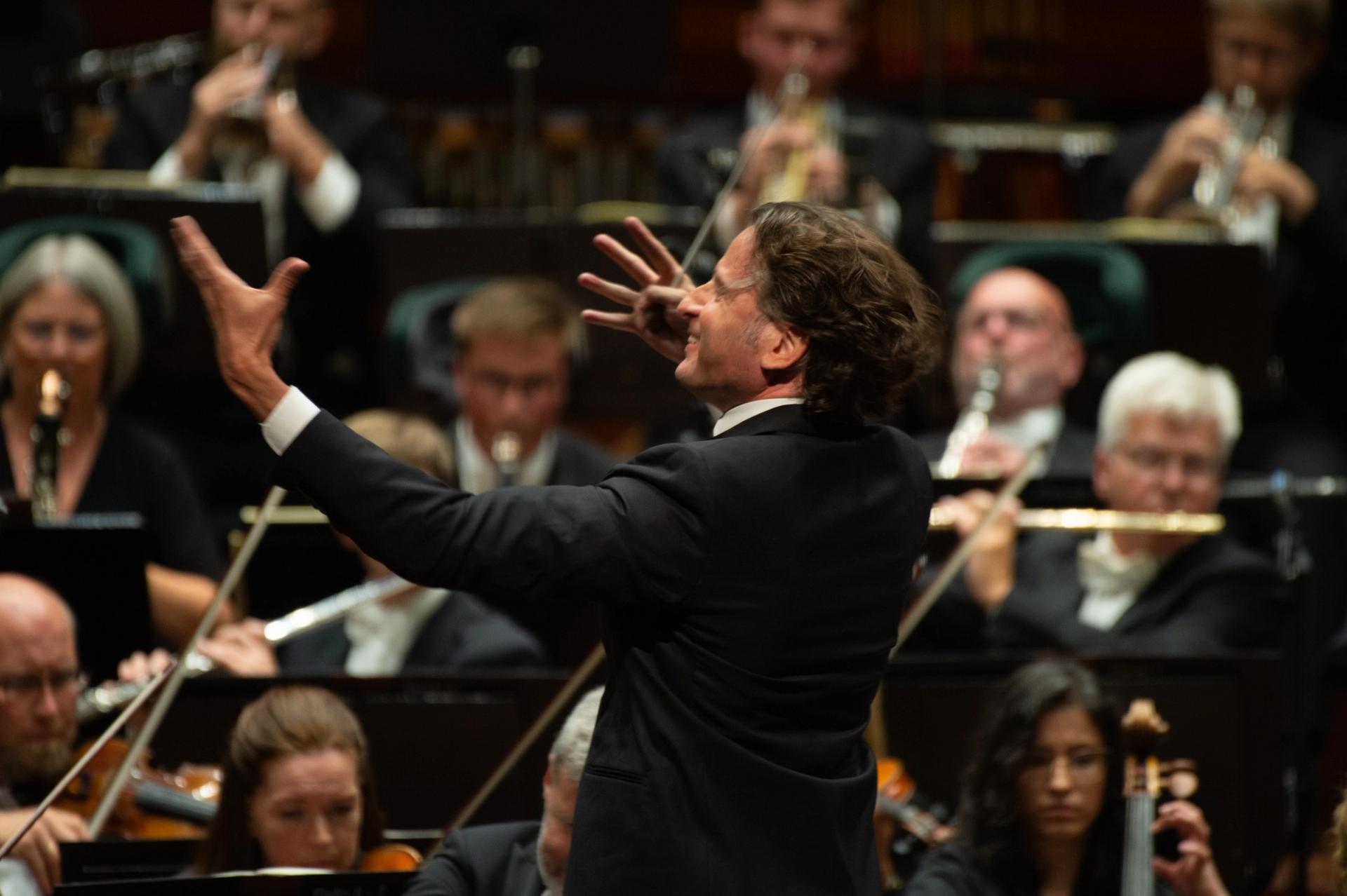 Dirigent leder Oslo-filharmonien under en konsert, med musikere i bakgrunnen.