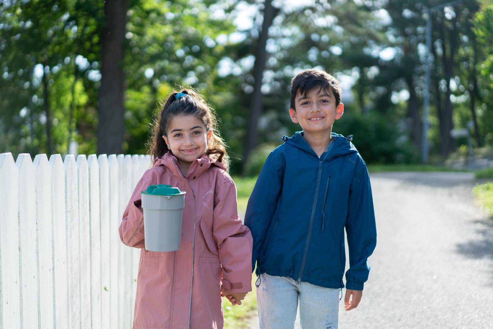 To barn smiler og holder hender, mens en av dem bærer en bøtte.