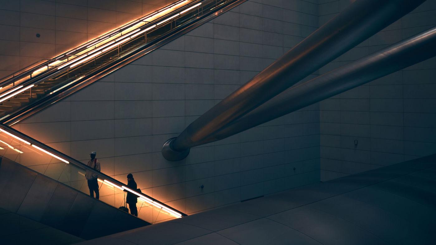 To mennesker på en rulletrapp opplyst av oransje lys i en ellers mørk bygning med store rør i bakgrunnen.
