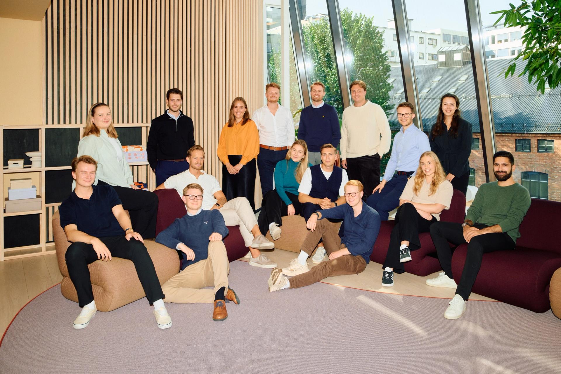 Gruppe mennesker poserer for et bilde foran et stort vindu, sittende og stående på en lilla matte og møbler.