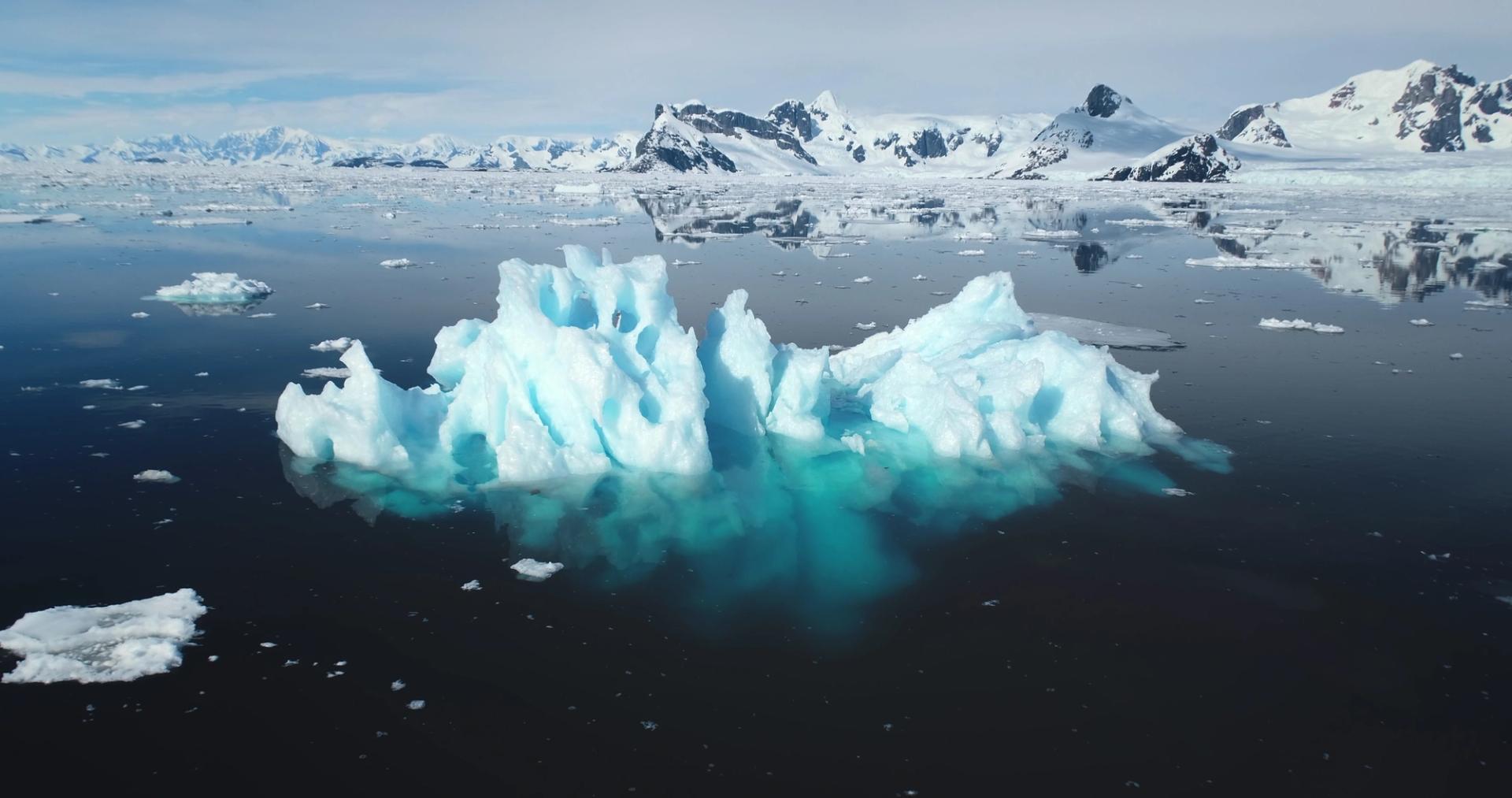 Et isblått isfjell i forgrunnen, med et isfylt hav og snøkledde fjell i bakgrunnen.