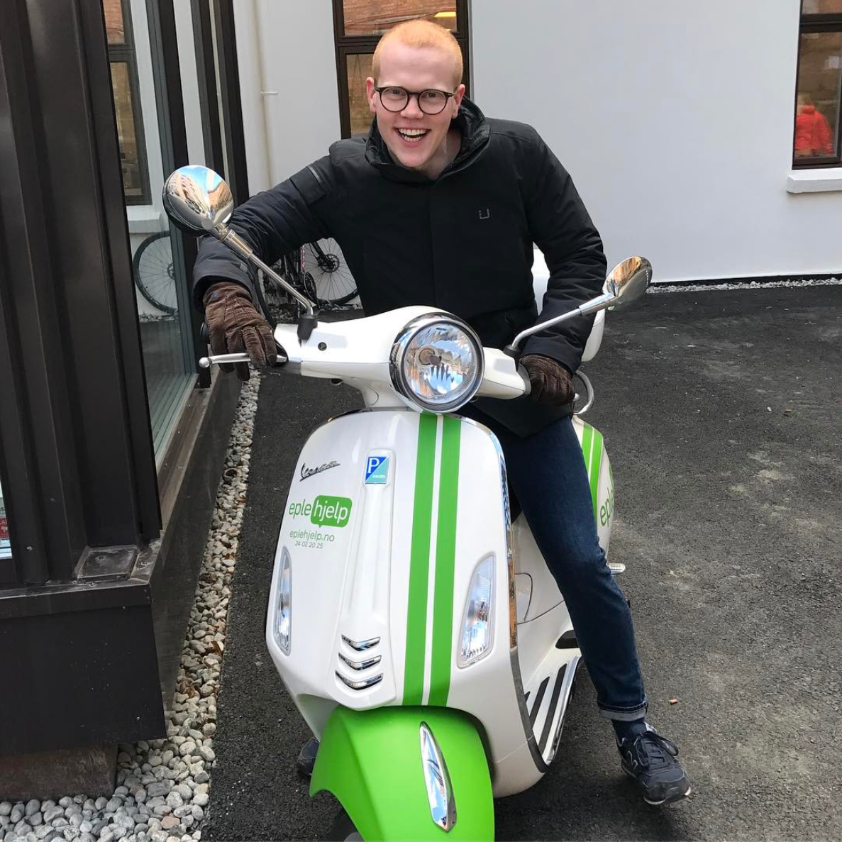 Glad mann sitter på en hvit Vespa-scooter med grønne striper, utenfor Handelshøyskolen BI.