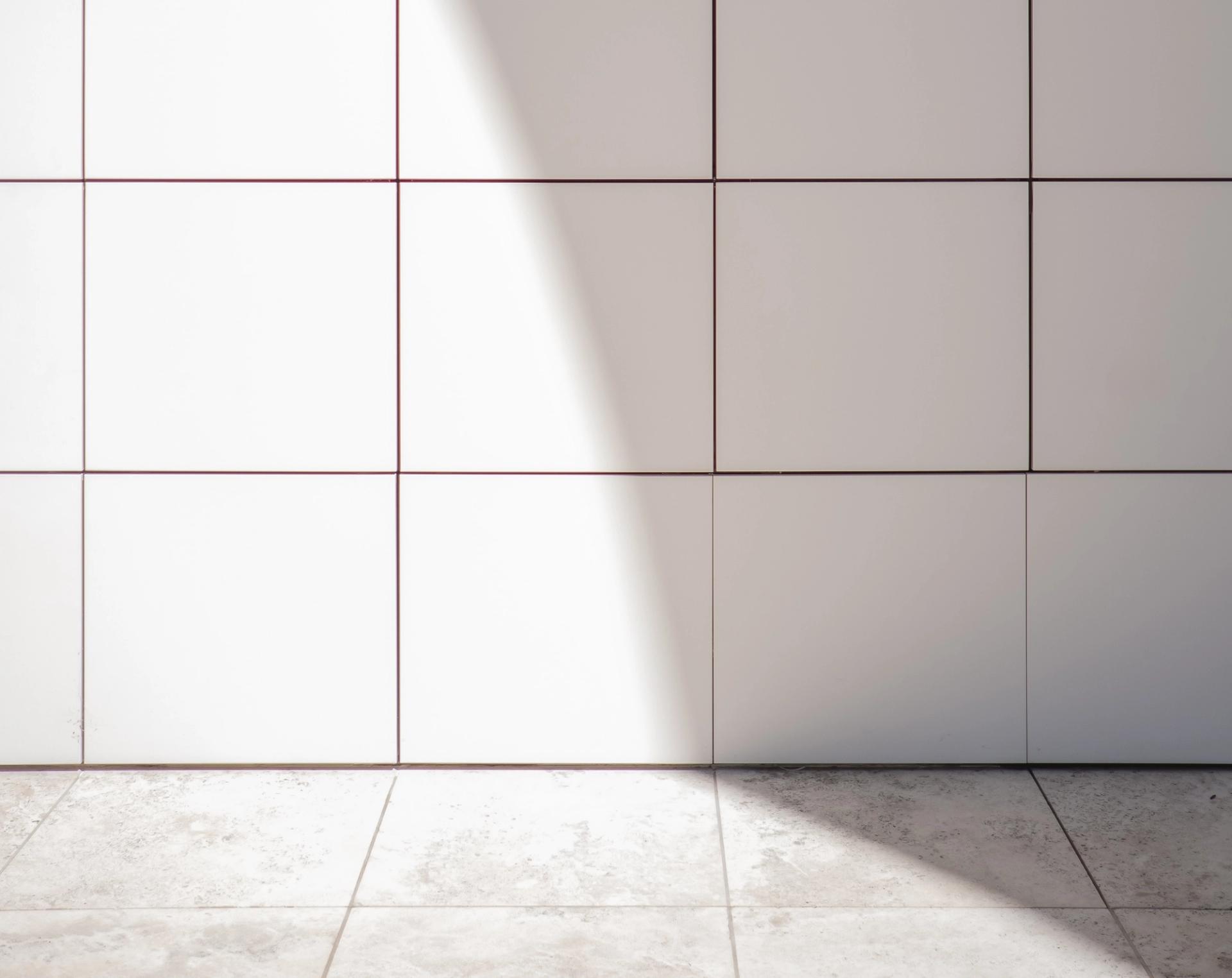 Lys vegg med hvite fliser og tynne røde fuger, og et lyst steingulv med solskinn på.