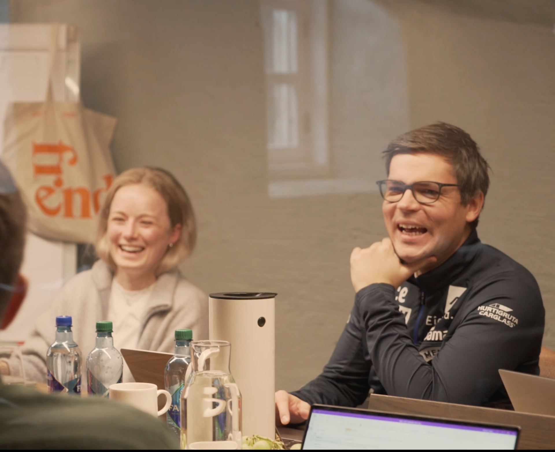 Møtedeltakere smiler og ler rundt et bord med flasker, krus, en laptop og en veske med ordet "friends".