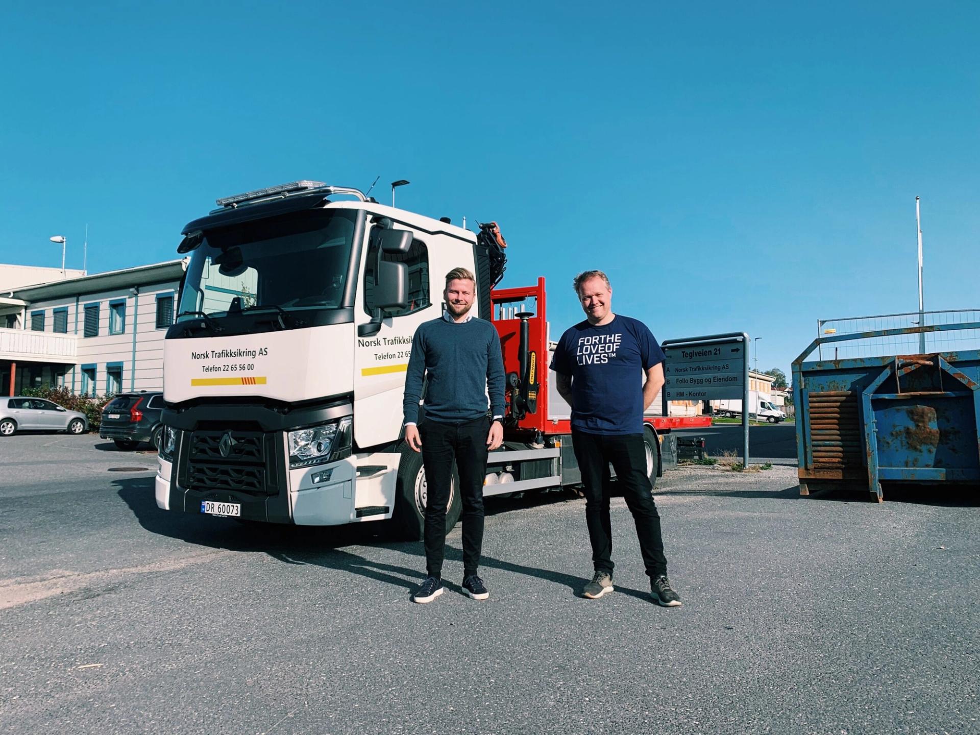 To menn smiler foran en hvit lastebil med "Norsk Trafikksikring AS" skrevet på siden.