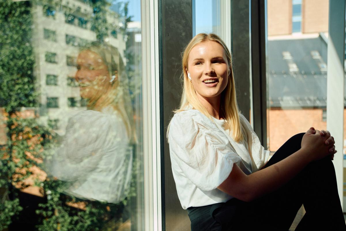 En smilende ung blond kvinne med ørepropper sitter ved et vindu med refleksjoner.
