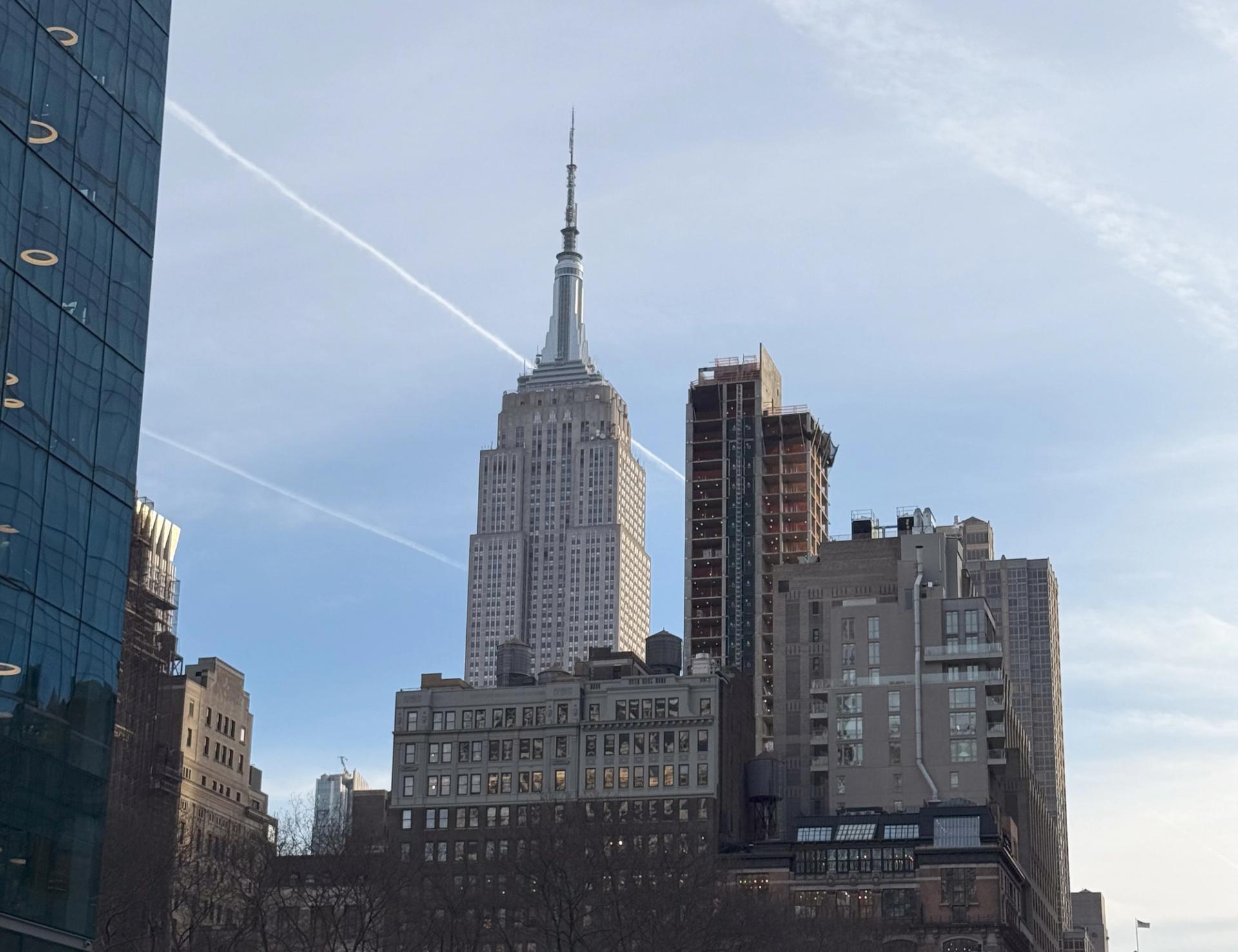 Bybildet av New York med Empire State Building i sentrum, omgitt av andre bygninger og en klar, lys himmel med kondensstriper.