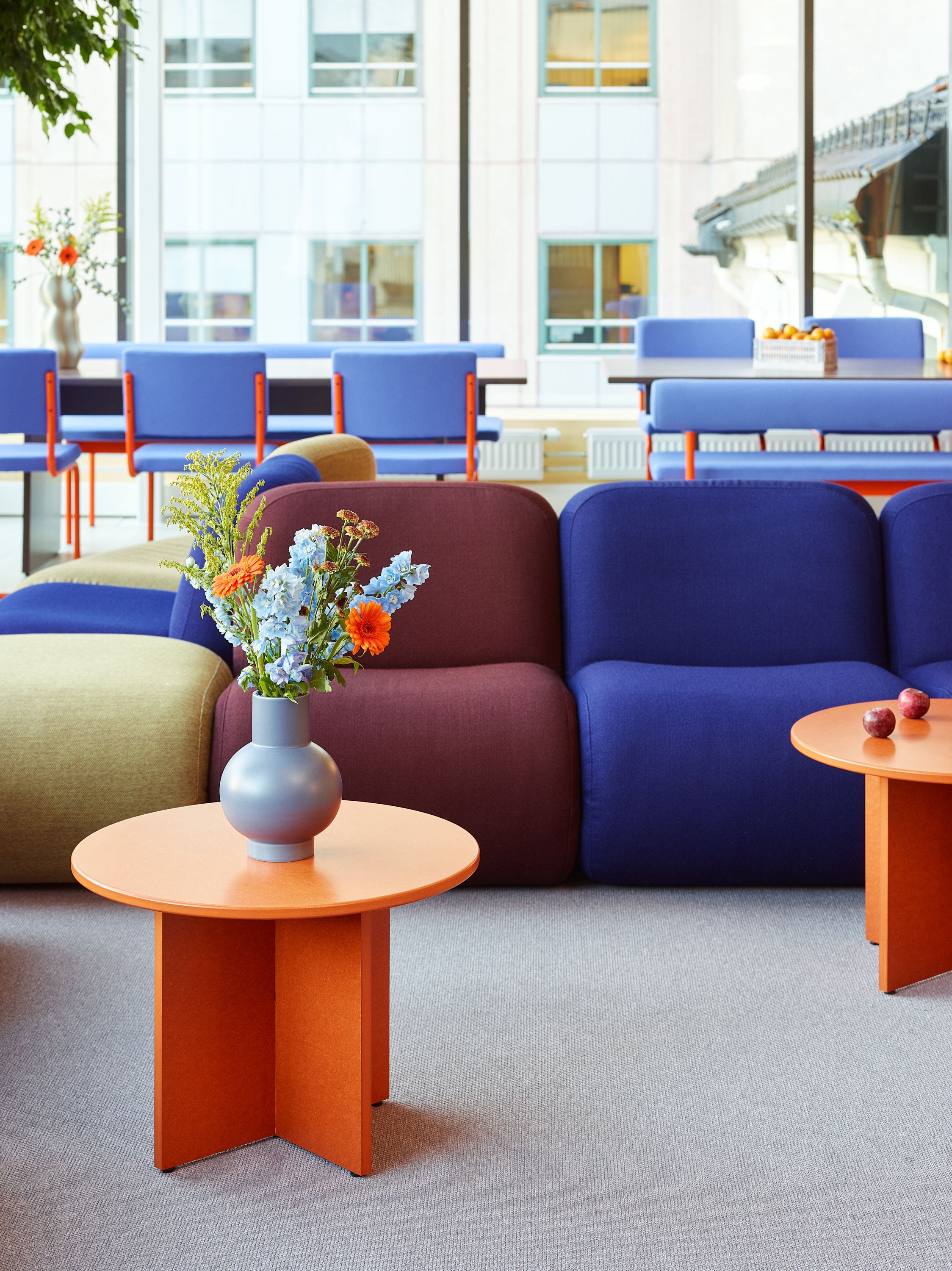 Fargerikt møblert kontorlandskap med blå, lilla og limegrønne sofaer, oransje bord og blomster i vaser.