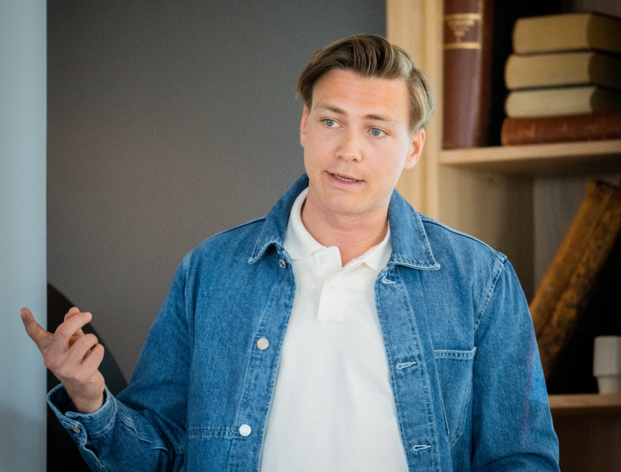 En ung mann i dongerijakke og hvit pologenser peker, med en bokhylle i bakgrunnen.