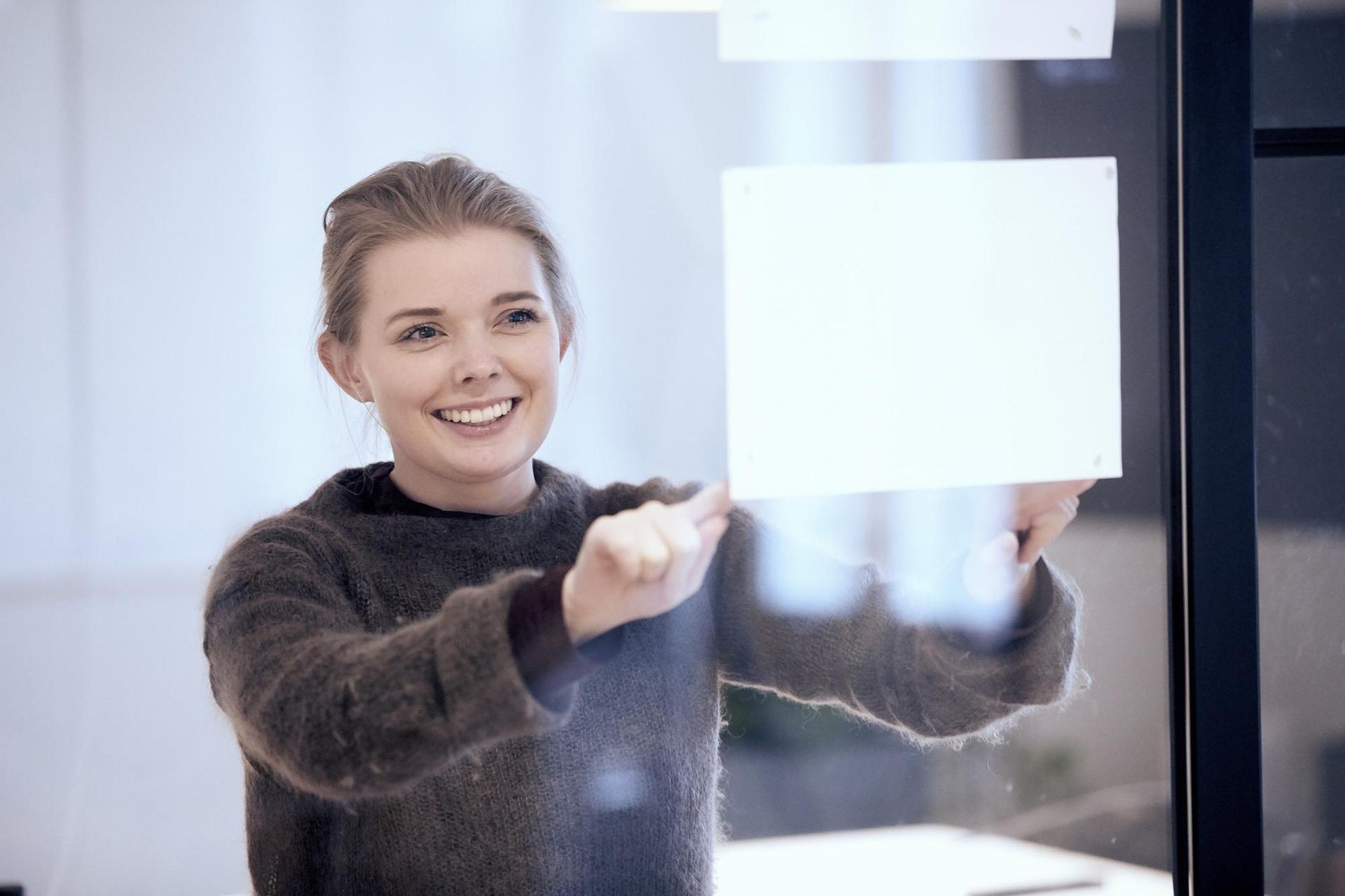 Smilende kvinne fester hvite lapper på en glassvegg, reflekterende lys i rommet.