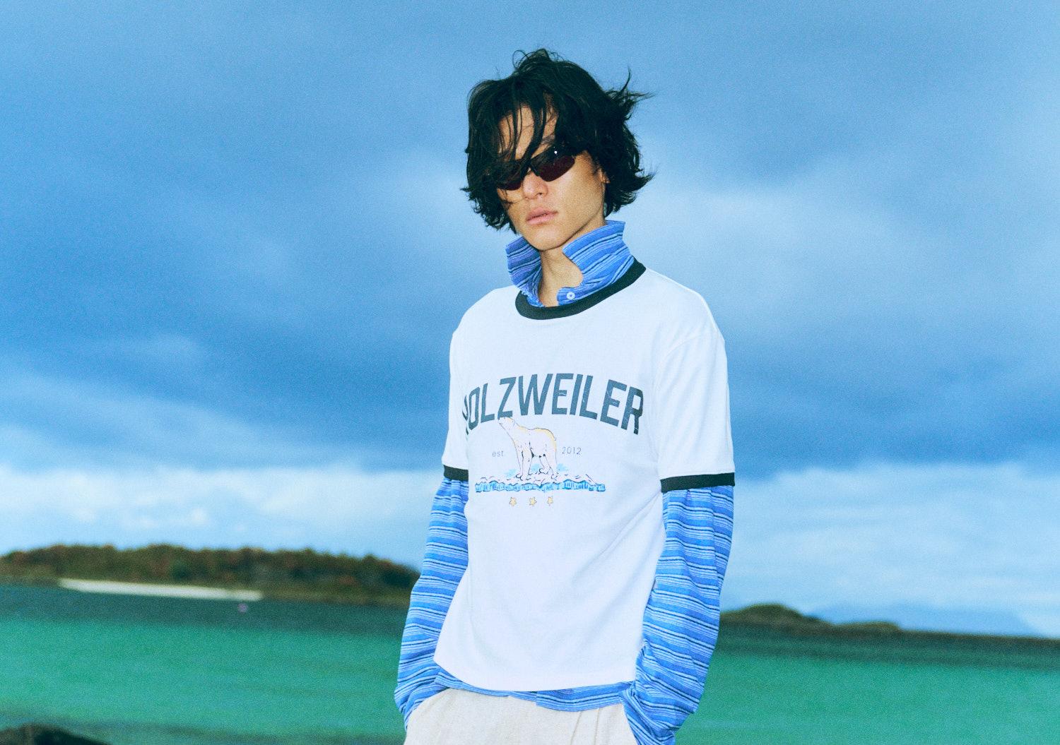 En ung person med solbriller og lag-på-lag klær står foran blå himmel og turkist hav.