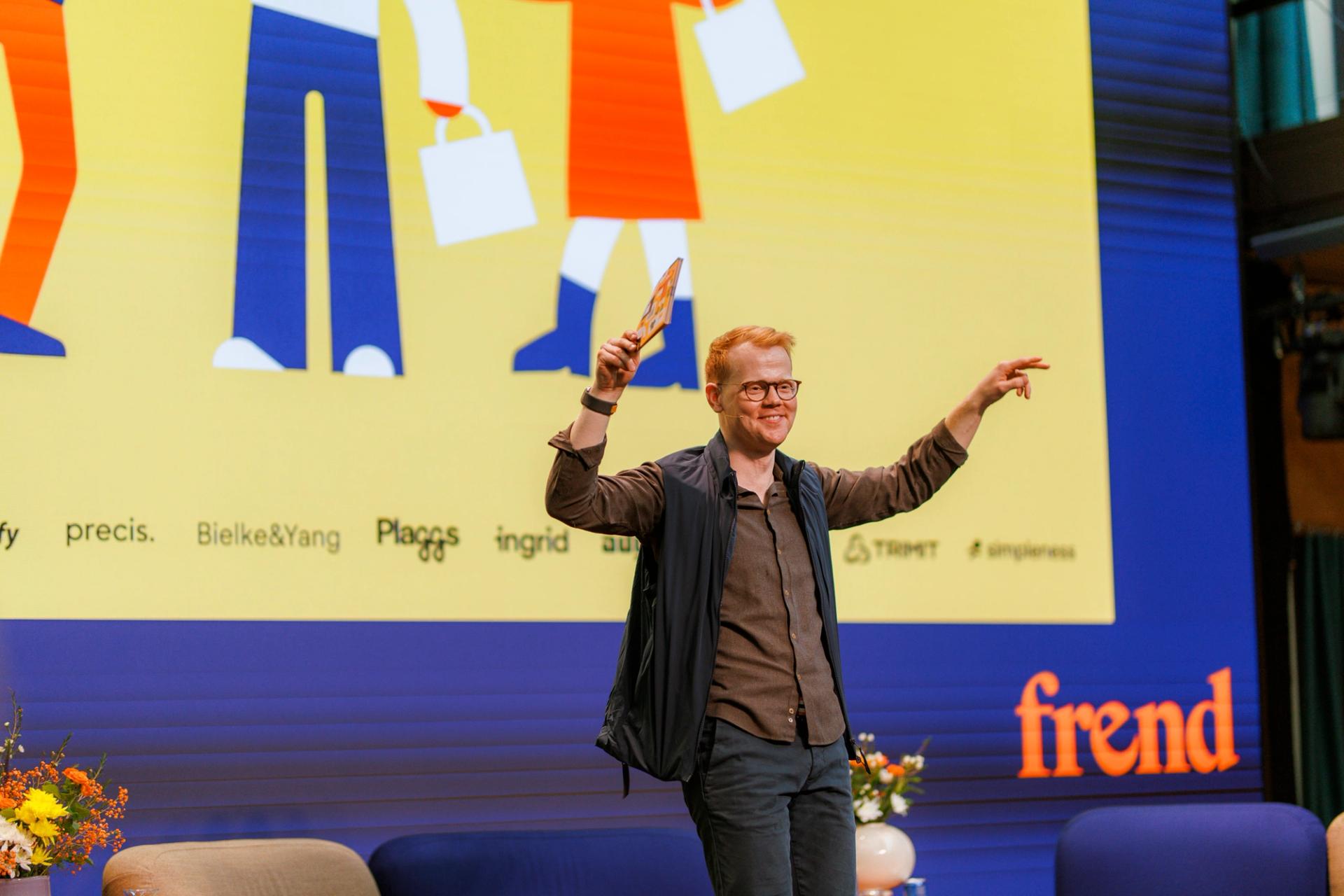 En smilende mann med briller på scenen, holder opp en gjenstand og peker mot publikum, foran et fargerikt bakteppe.