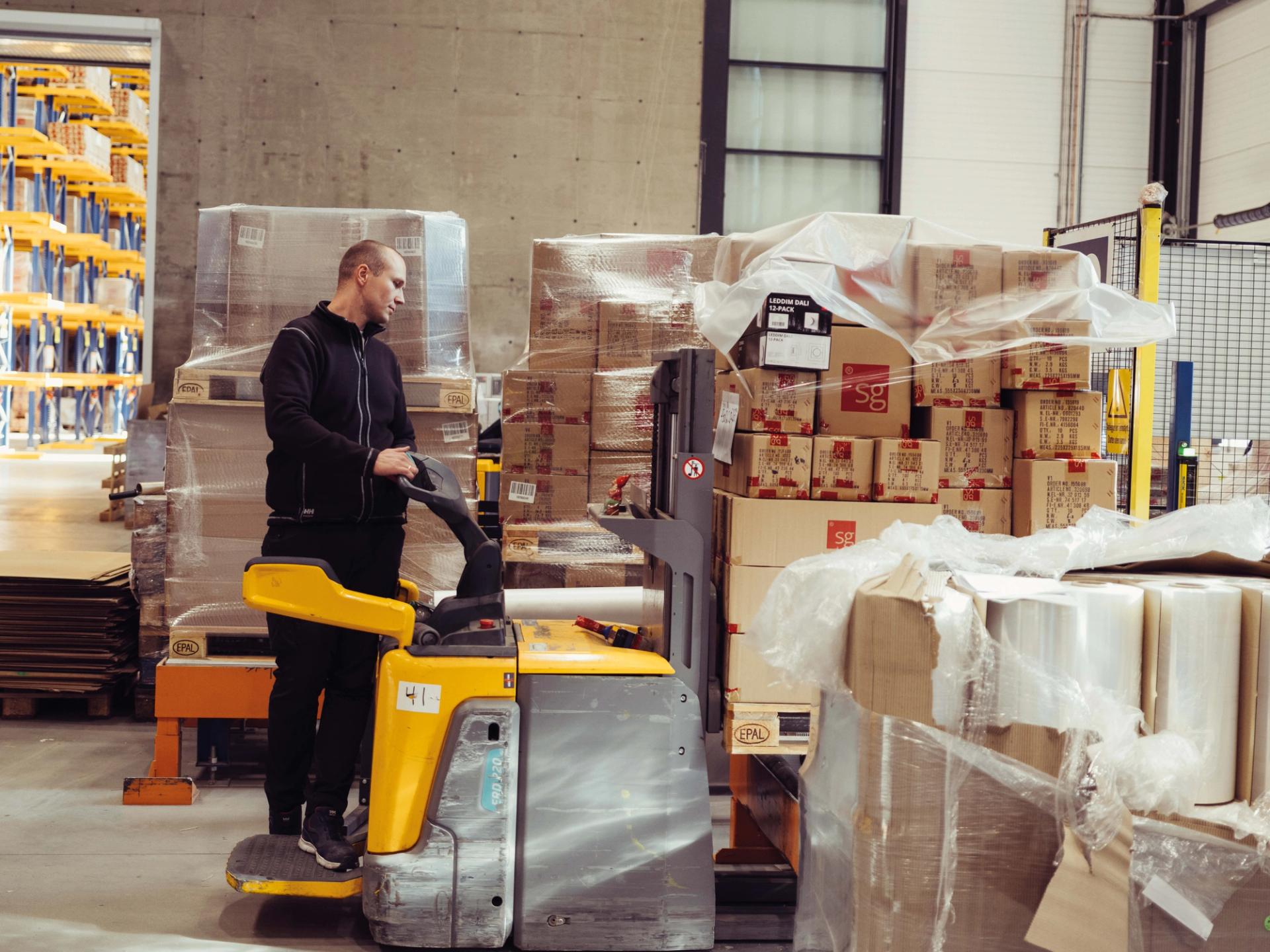 Mann på jekketralle i et lager, omgitt av paller med esker innpakket i plast.