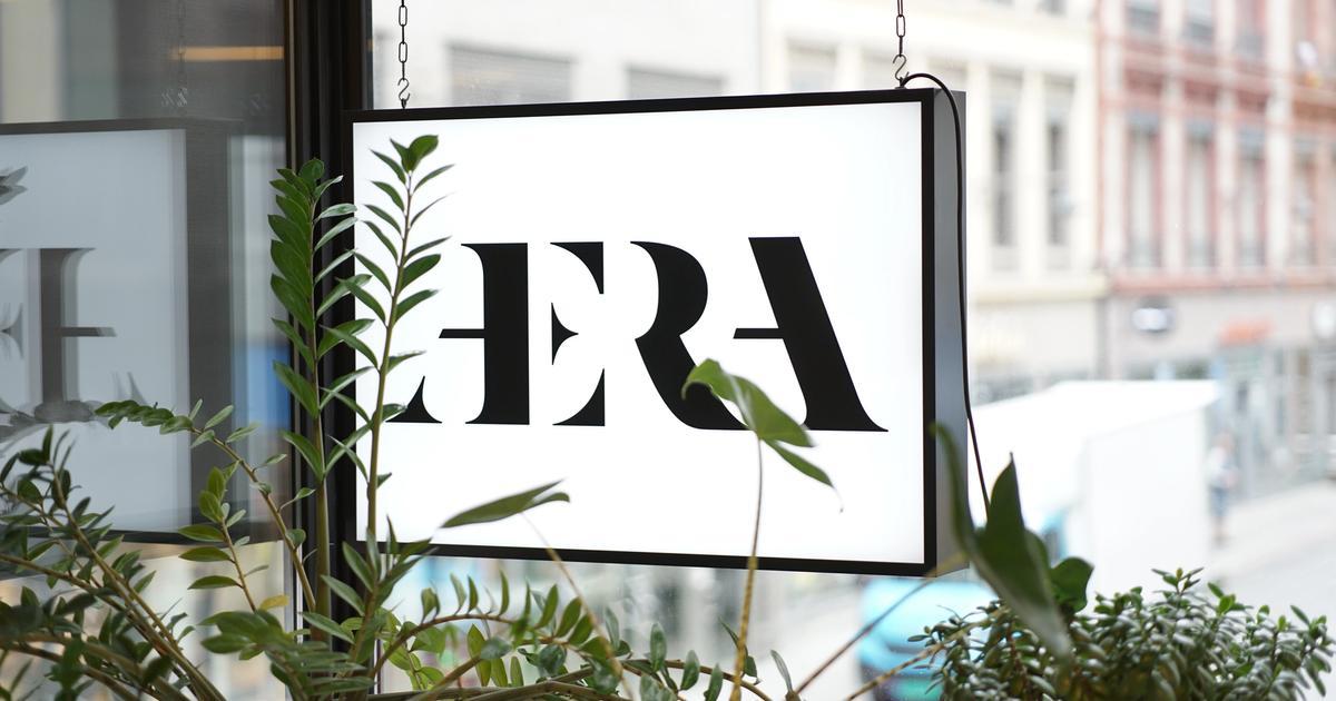 Lysskilt med "ÆRA" skrevet i svart skrift på hvit bakgrunn, henger i et vindu med planter i forgrunnen og bygning i bakgrunnen.