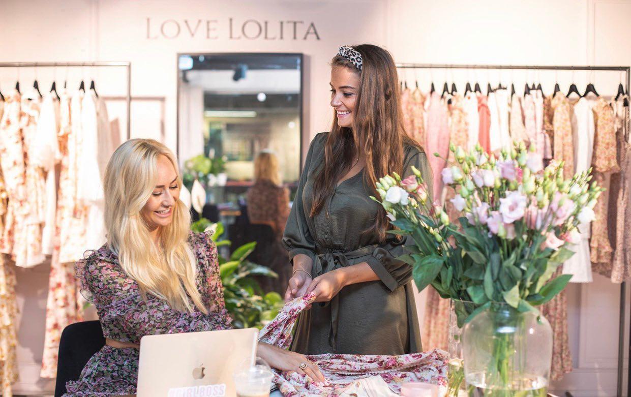 To kvinner smiler mens den ene bretter et plagg og den andre jobber på en laptop, med klær hengende i bakgrunnen.