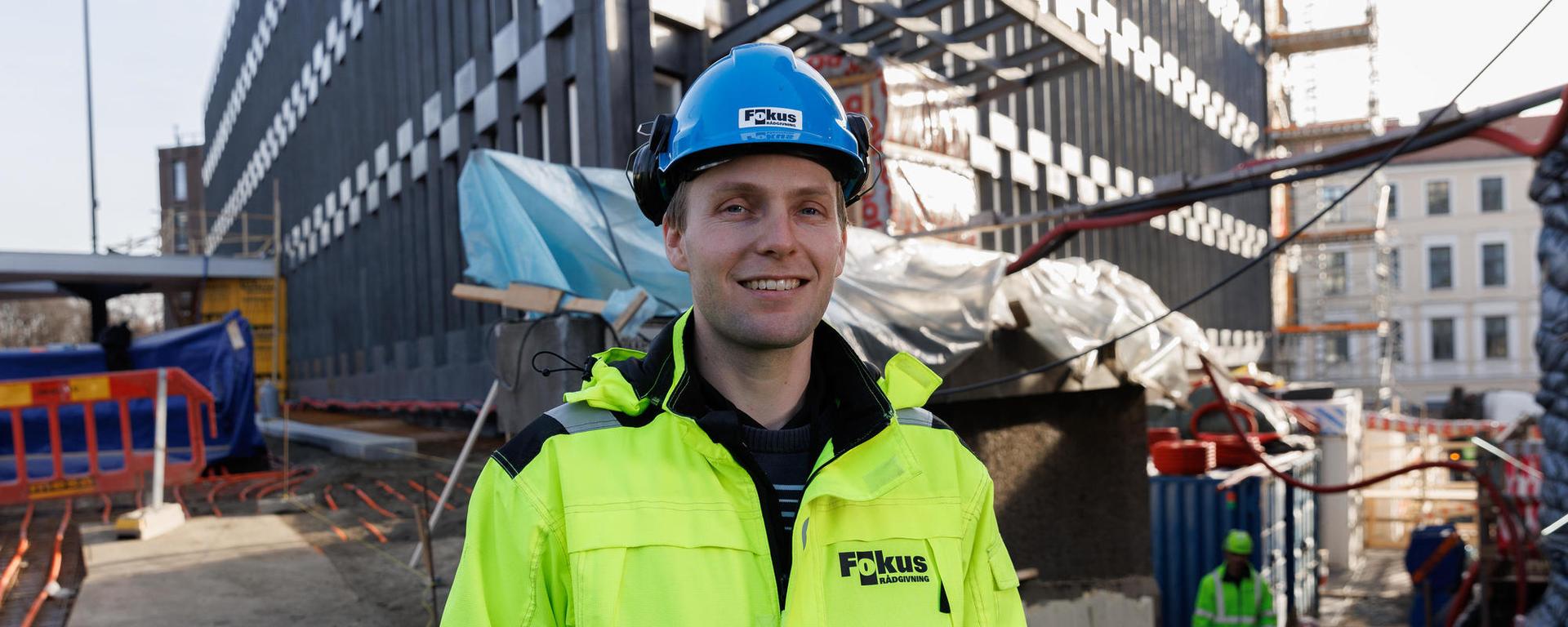 En smilende mann i gul refleksjakke og blå hjelm på en byggeplass.
