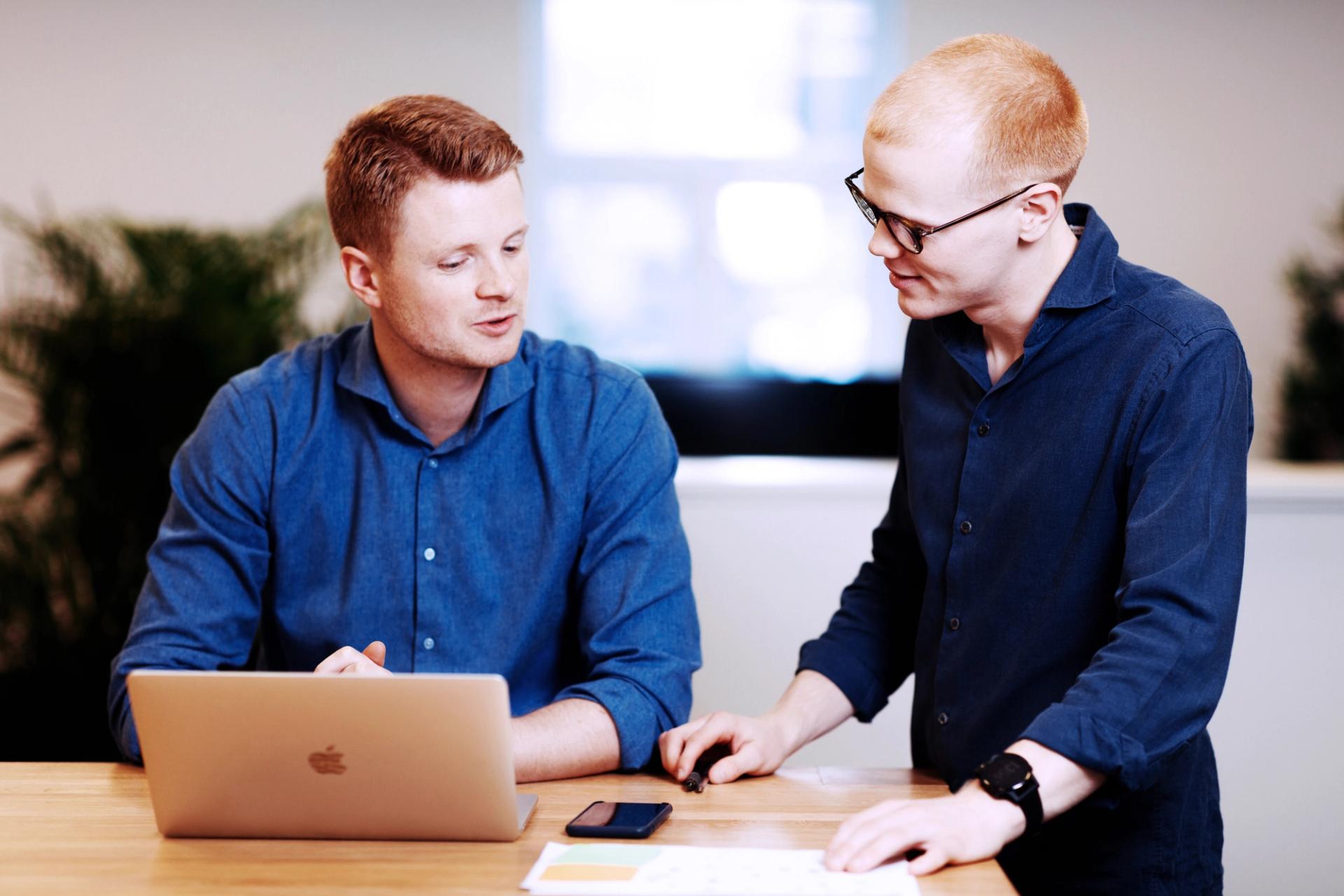 To menn i blå skjorter jobber sammen ved et bord med en bærbar PC, mobiltelefon og papirer.