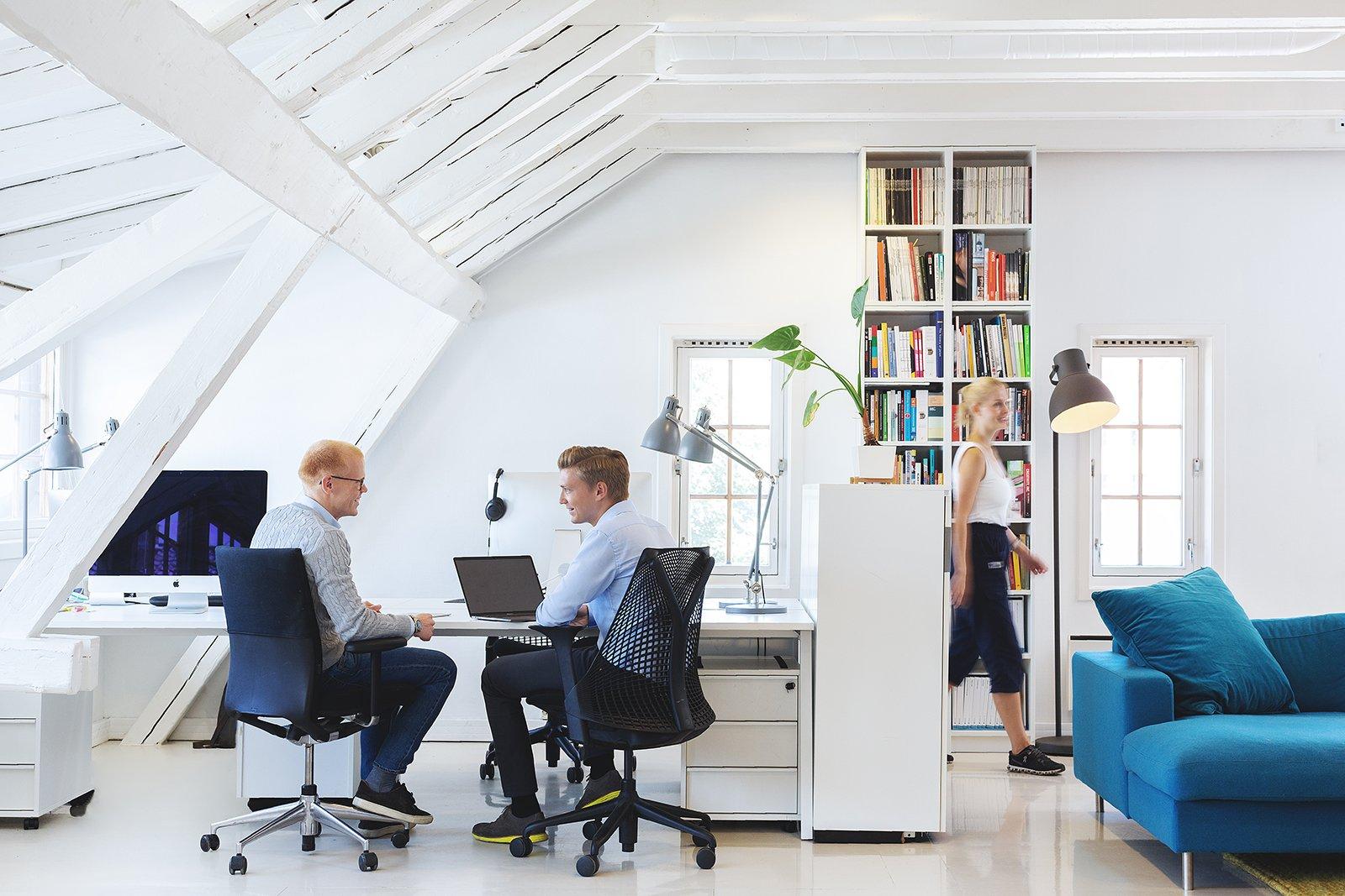 Tre kolleger jobber og går i et lyst, moderne kontorlokale med hvite vegger, takbjelker og en blå sofa.