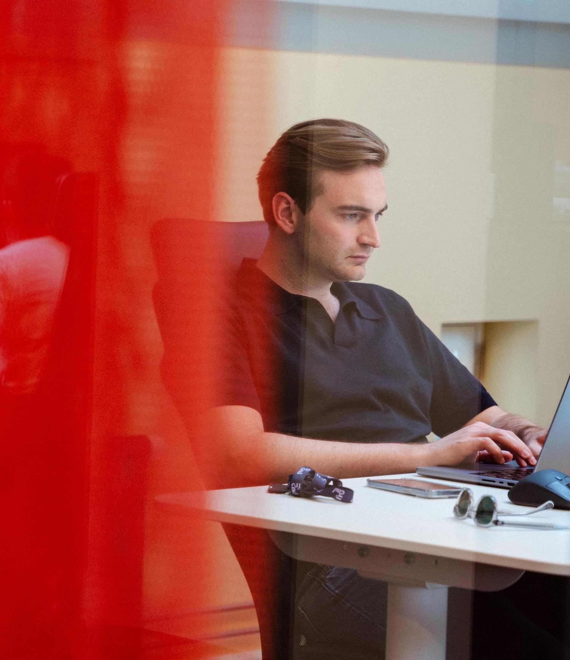 En mann jobber på en laptop, delvis skjult bak en rød transparent skillevegg. En annen person jobber i bakgrunnen.