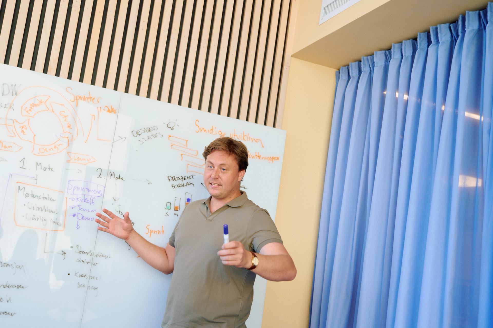 Mann i polotrøye presenterer et smidig utviklingsdiagram på en tavle, med blå gardiner i bakgrunnen.