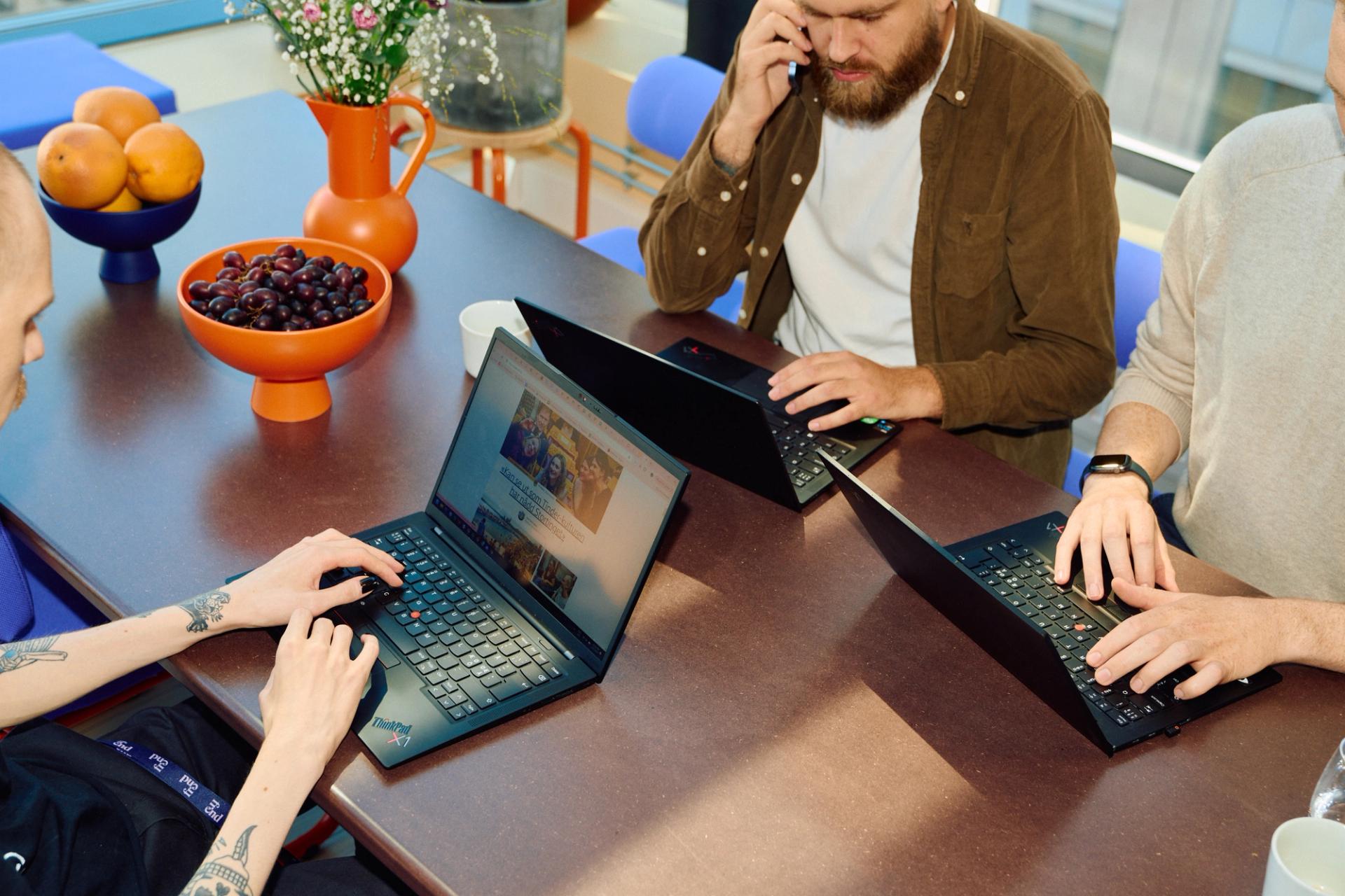 Tre personer jobber på bærbare datamaskiner rundt et bord med frukt og blomster i lyse omgivelser.