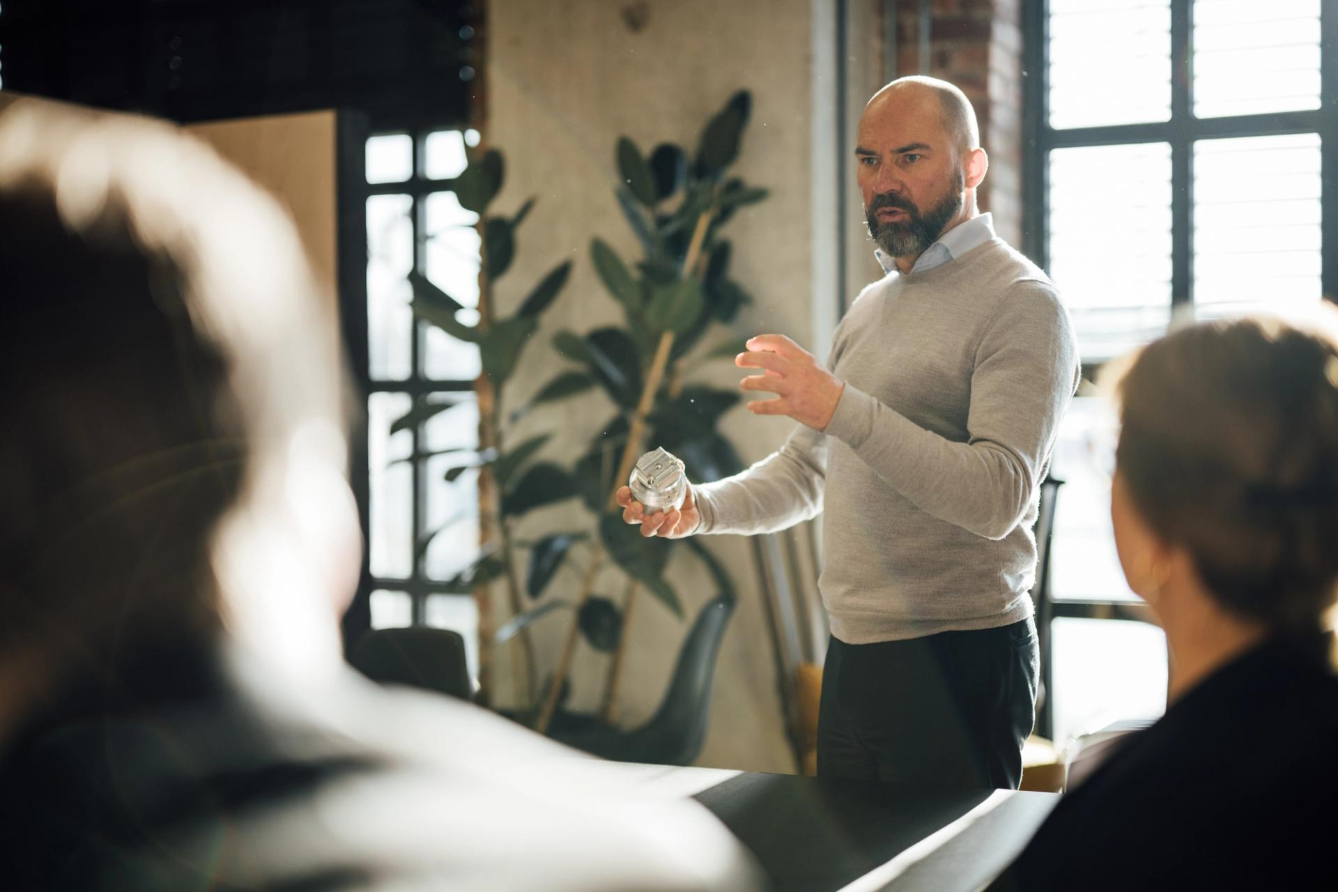 En mann presenterer for to uskarpe personer, holder et lite metallobjekt i den ene hånden.