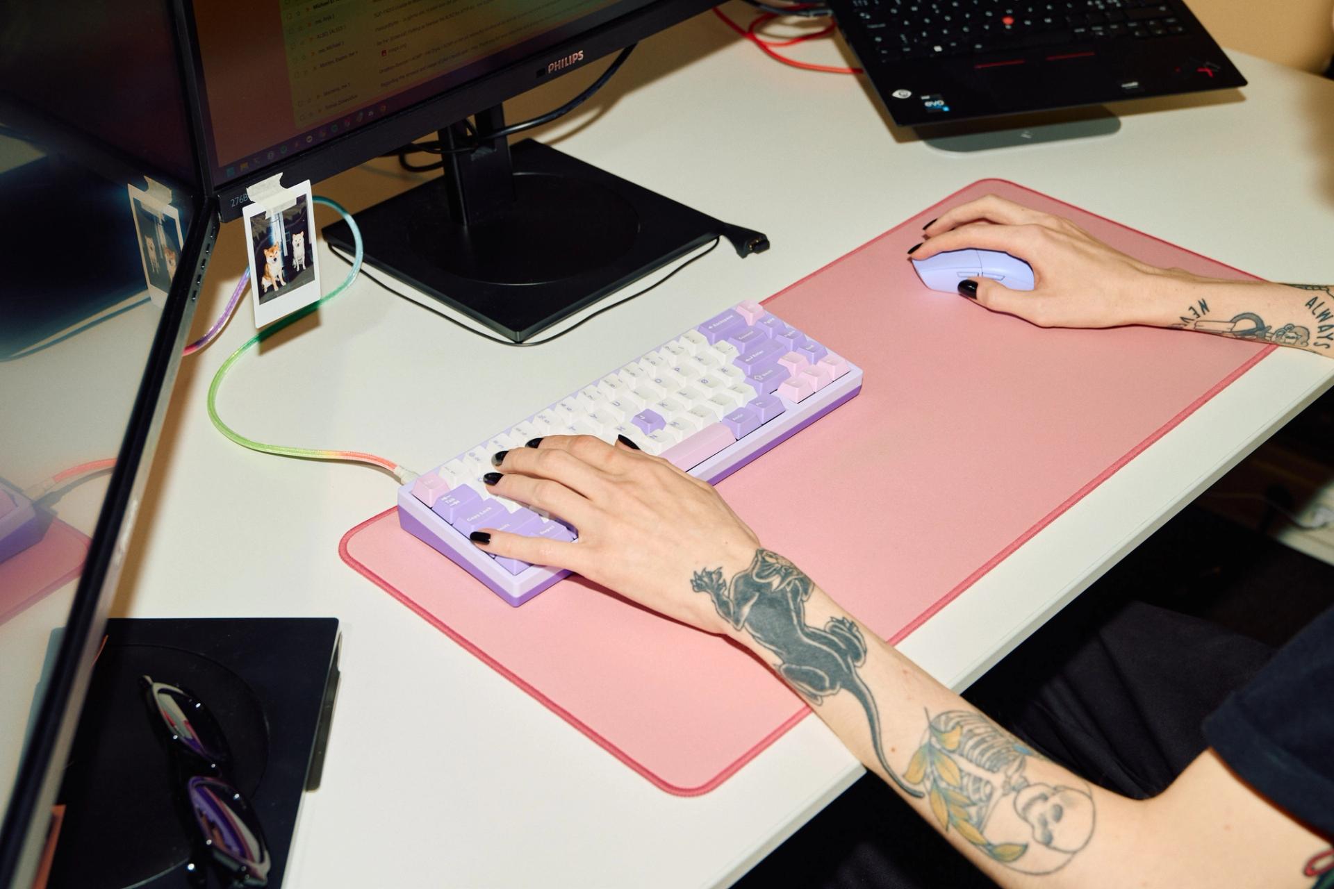 En person med tatoveringer bruker et lilla tastatur og en lilla mus på en rosa musematte ved en datamaskin.