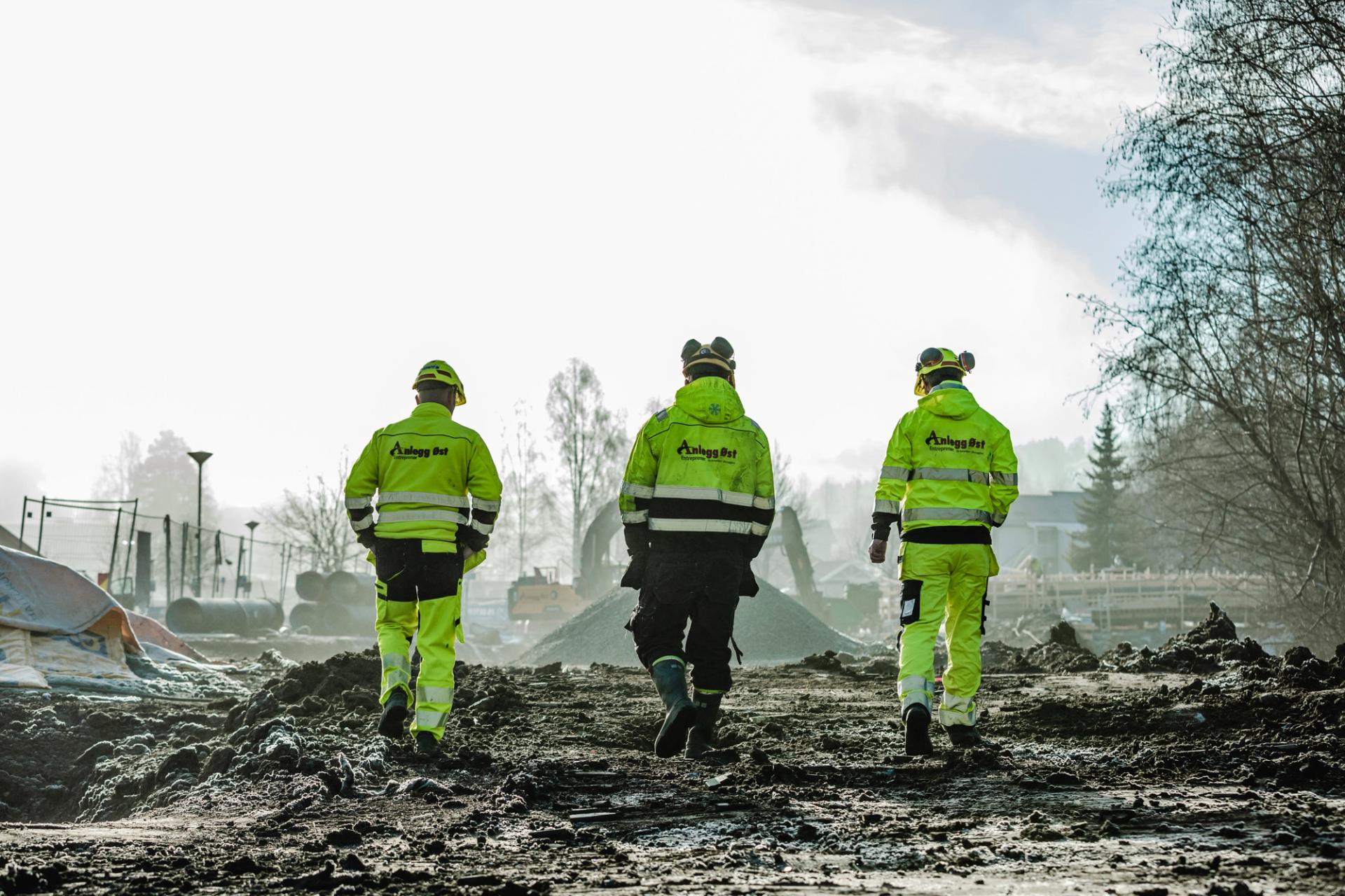 Tre arbeidere i gul refleksbekledning går over et gjørmete, frossent anleggsområde.