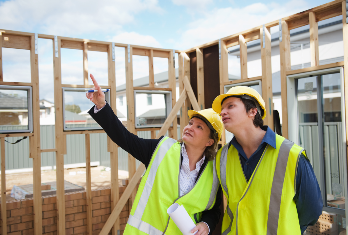 To bygningsarbeidere i refleksvester og hjelm peker og ser opp på en bygningskonstruksjon.