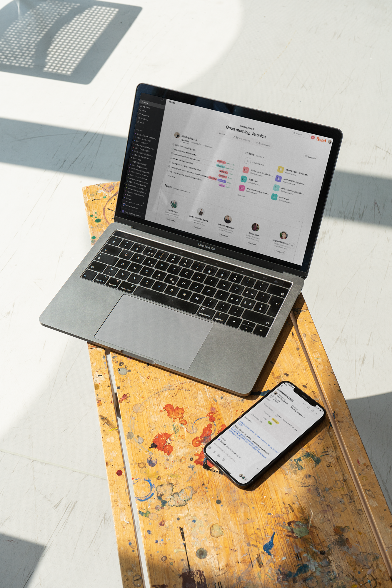 En MacBook Pro og en iPhone viser prosjektoversikter på en flekkete trebordsflate.