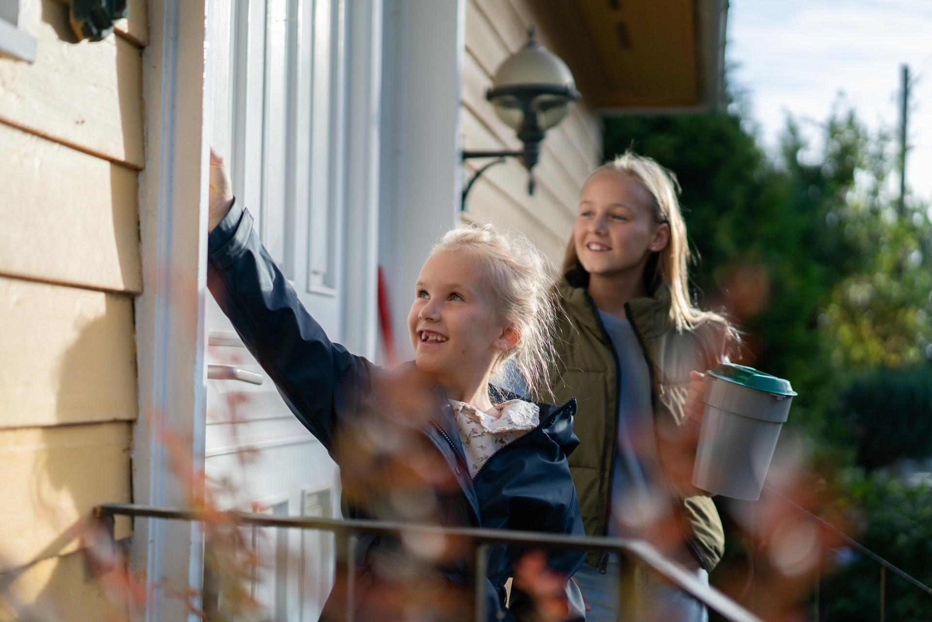 To jenter med en innsamlingsbøsse står ved en dør og smiler.
