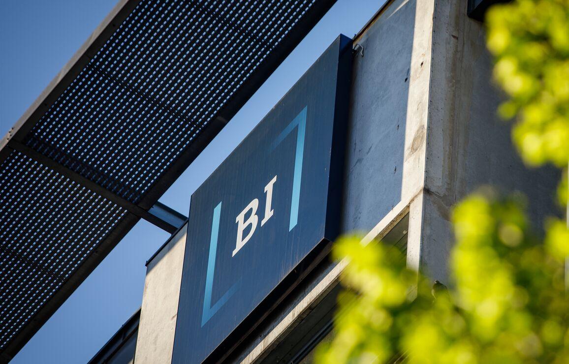 Skilt med "BI" på en bygning, sett gjennom grønne blader mot en blå himmel.