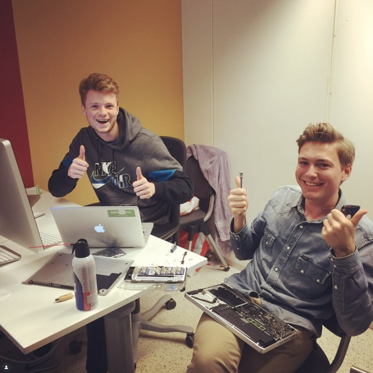 To karer smiler og viser tommel opp; den ene jobber på en bærbar PC, den andre holder en demontert bærbar PC.