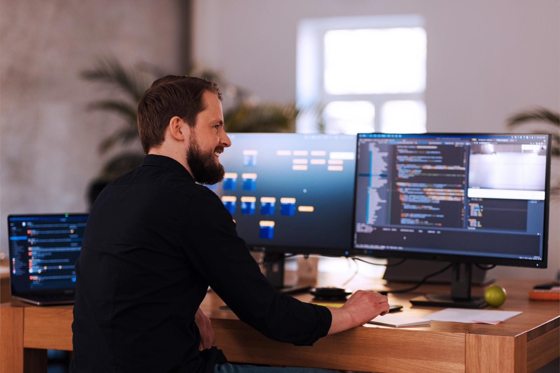 En smilende skjeggete mann i svart skjorte sitter ved et skrivebord med tre dataskjermer og en bærbar datamaskin.