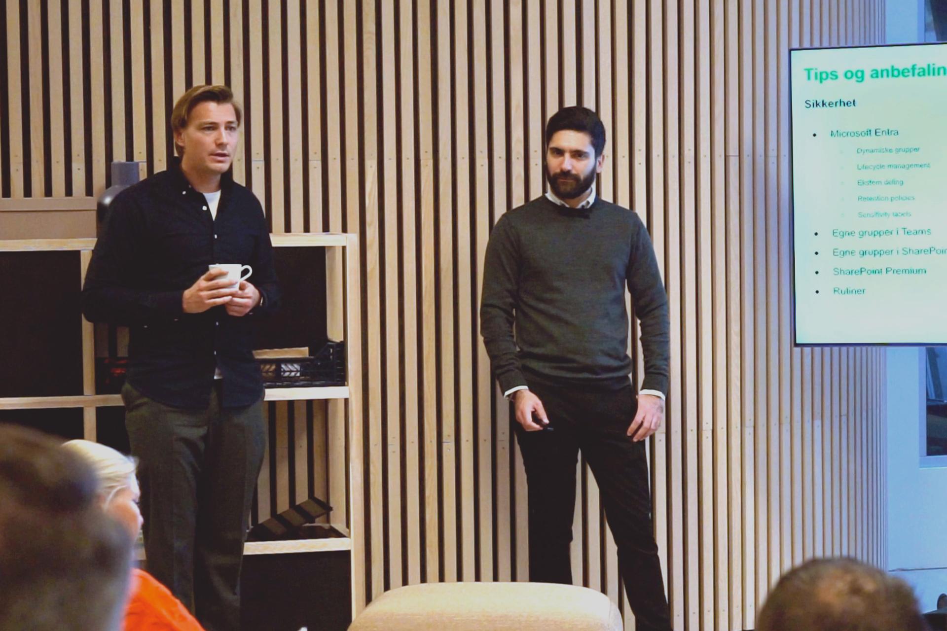 To menn presenterer på et seminar om SharePoint og Teams. En mann holder en kopp, den andre står med hendene i lommene.