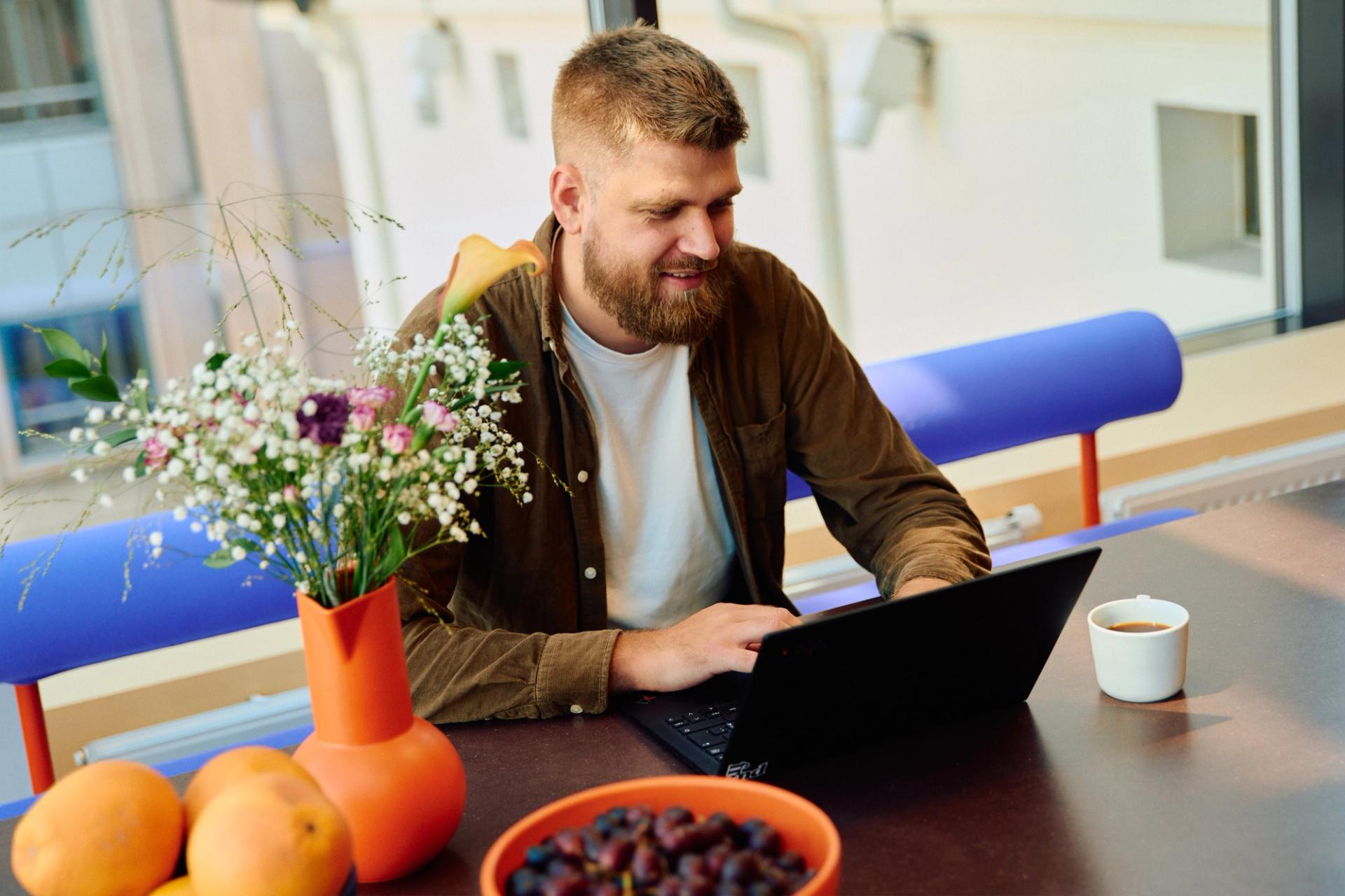 En smilende mann jobber på en laptop ved et bord med frukt, kaffe og blomster.