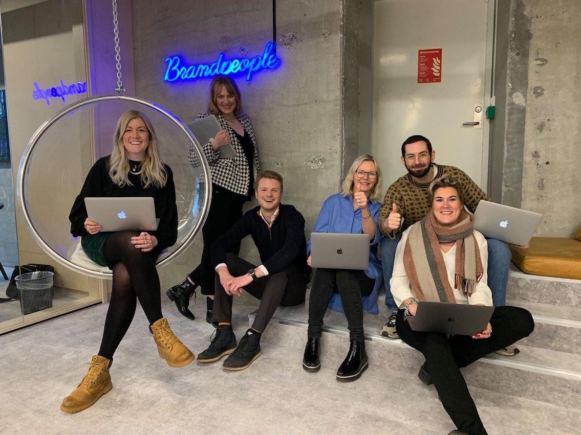 Seks smilende kolleger poserer med bærbare PC-er foran en neonbelyst vegg med "Brandpeople".