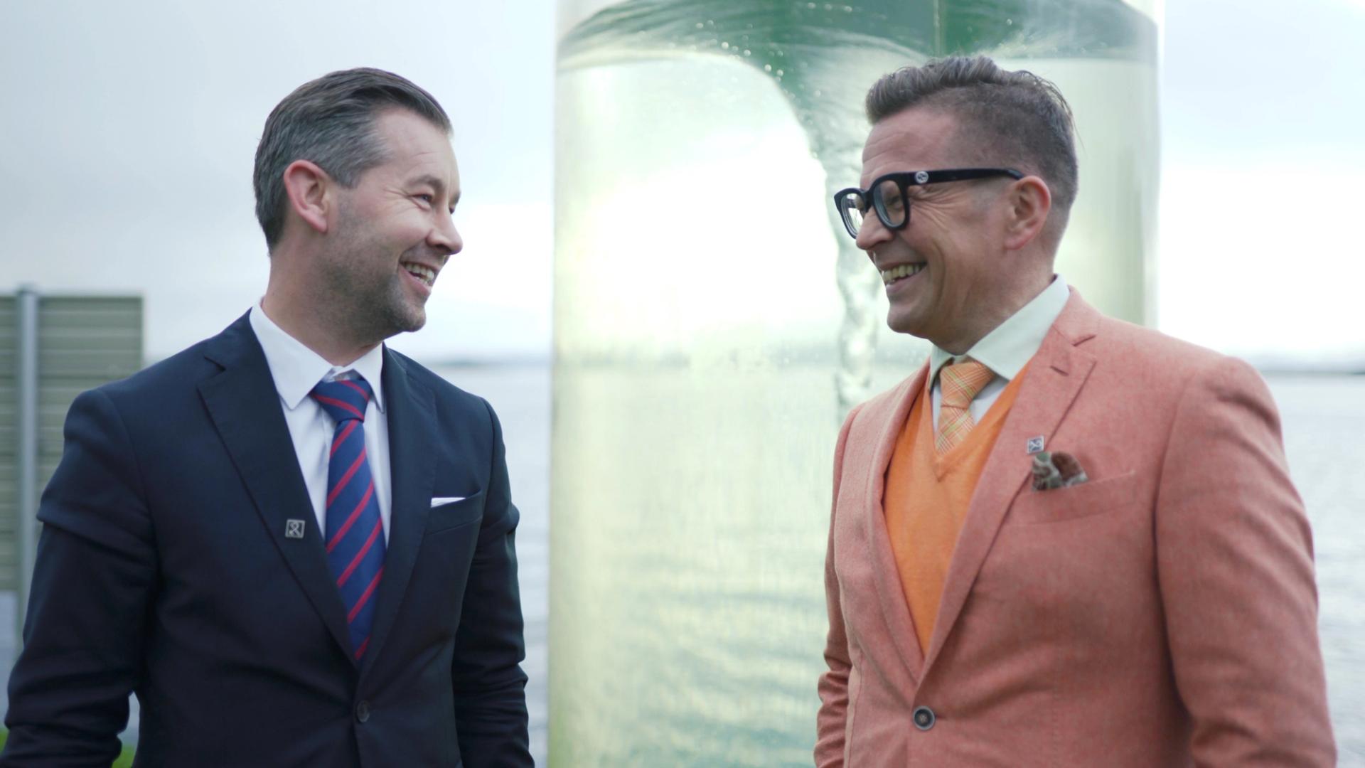 To menn i dress og blazer smiler mot hverandre foran et vannmagasin med en virvel.
