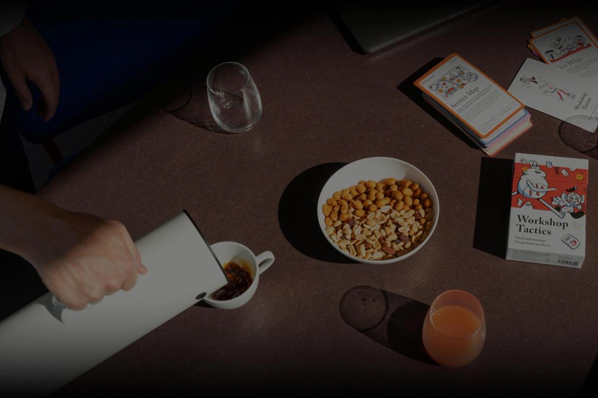 En person heller kaffe fra en hvit termos på et bord med en skål nøtter, juice og kortstokken "Workshop Tactics".