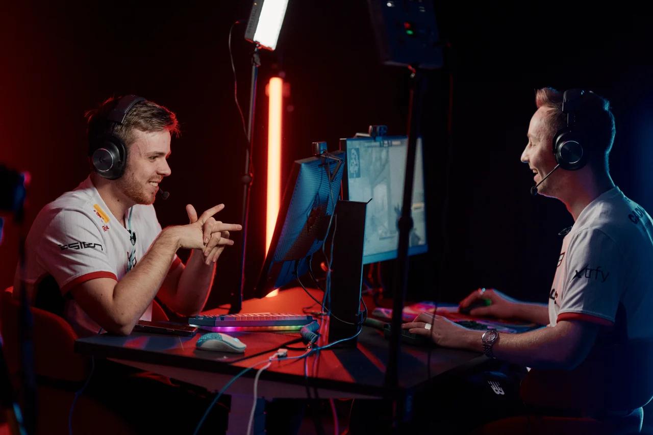 To menn med headset sitter foran dataskjermer og snakker, med gamingutstyr og rødt og blått lys rundt.