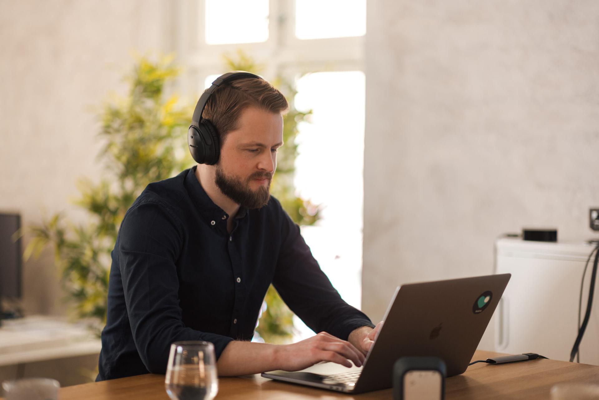Mann med skjegg og hodetelefoner jobber på en bærbar PC ved et trebord.