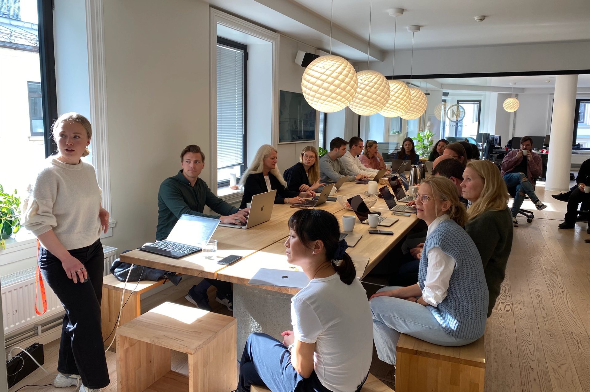 Folk sitter rundt et stort bord med bærbare datamaskiner og drikker kaffe, mens sollys skinner inn gjennom vinduene.